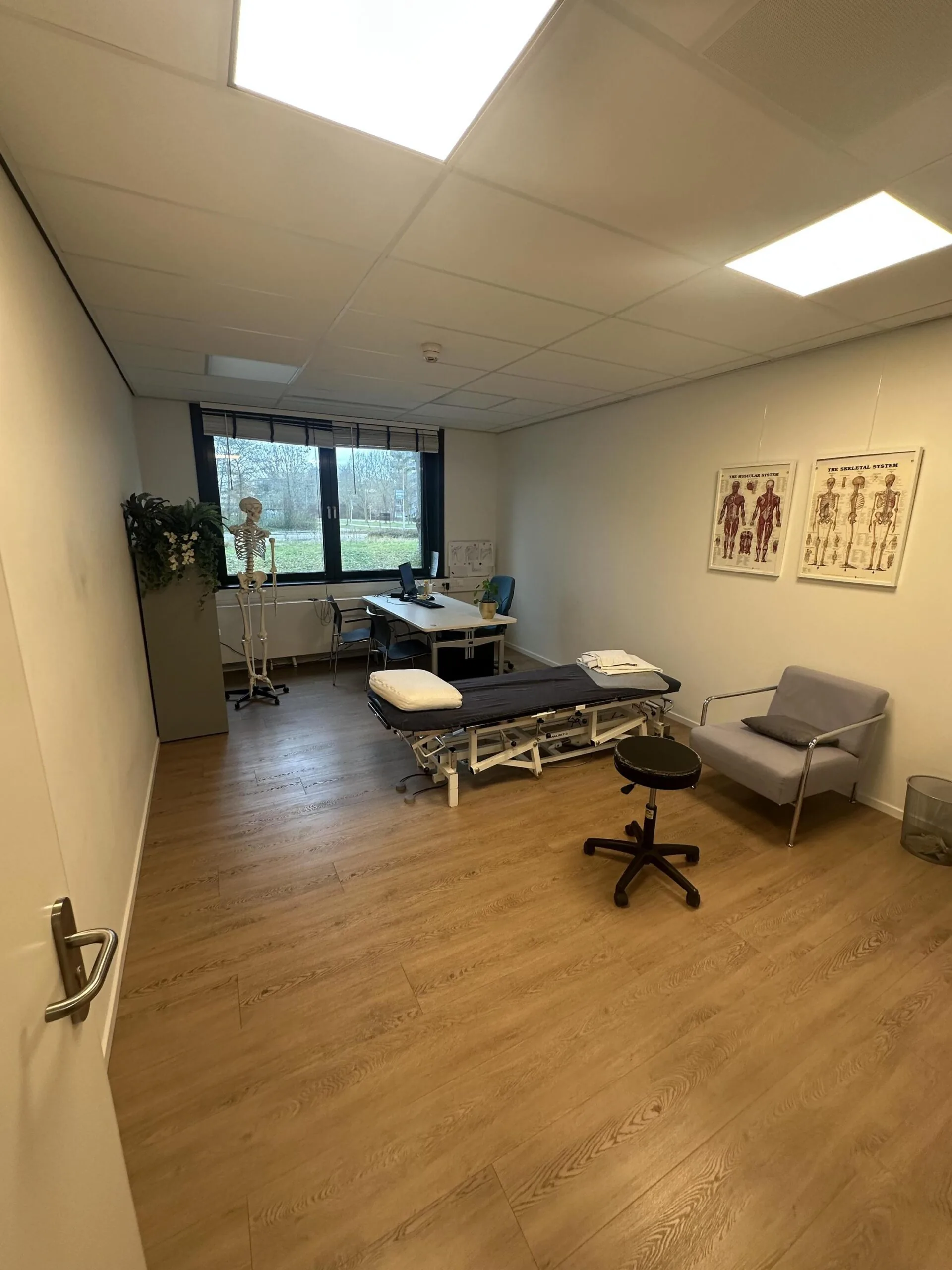 A modern medical or physiotherapy consultation room at Veteranenlaan, featuring an examination table, anatomical posters, and a desk by the window.