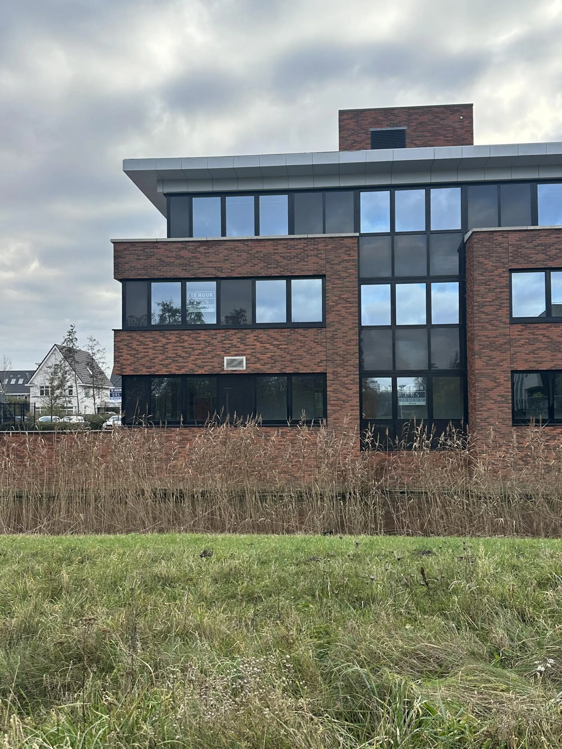 Modern brick office building on Veteranenlaan with a "For Rent" sign in the window, viewed across a grassy area and reeds.