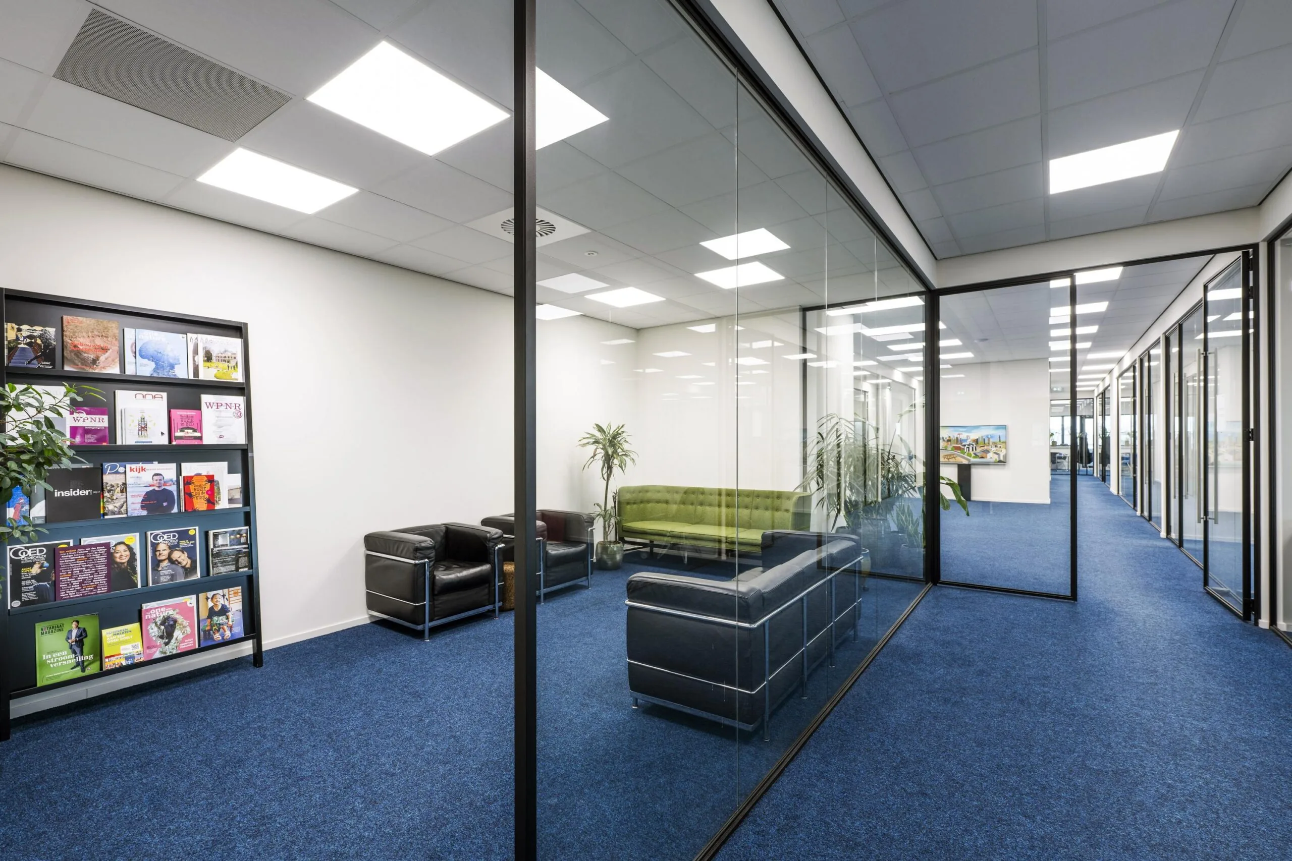 Modern office hallway on Dynamostraat with glass walls, blue carpet, and a waiting area furnished with black and green seating.