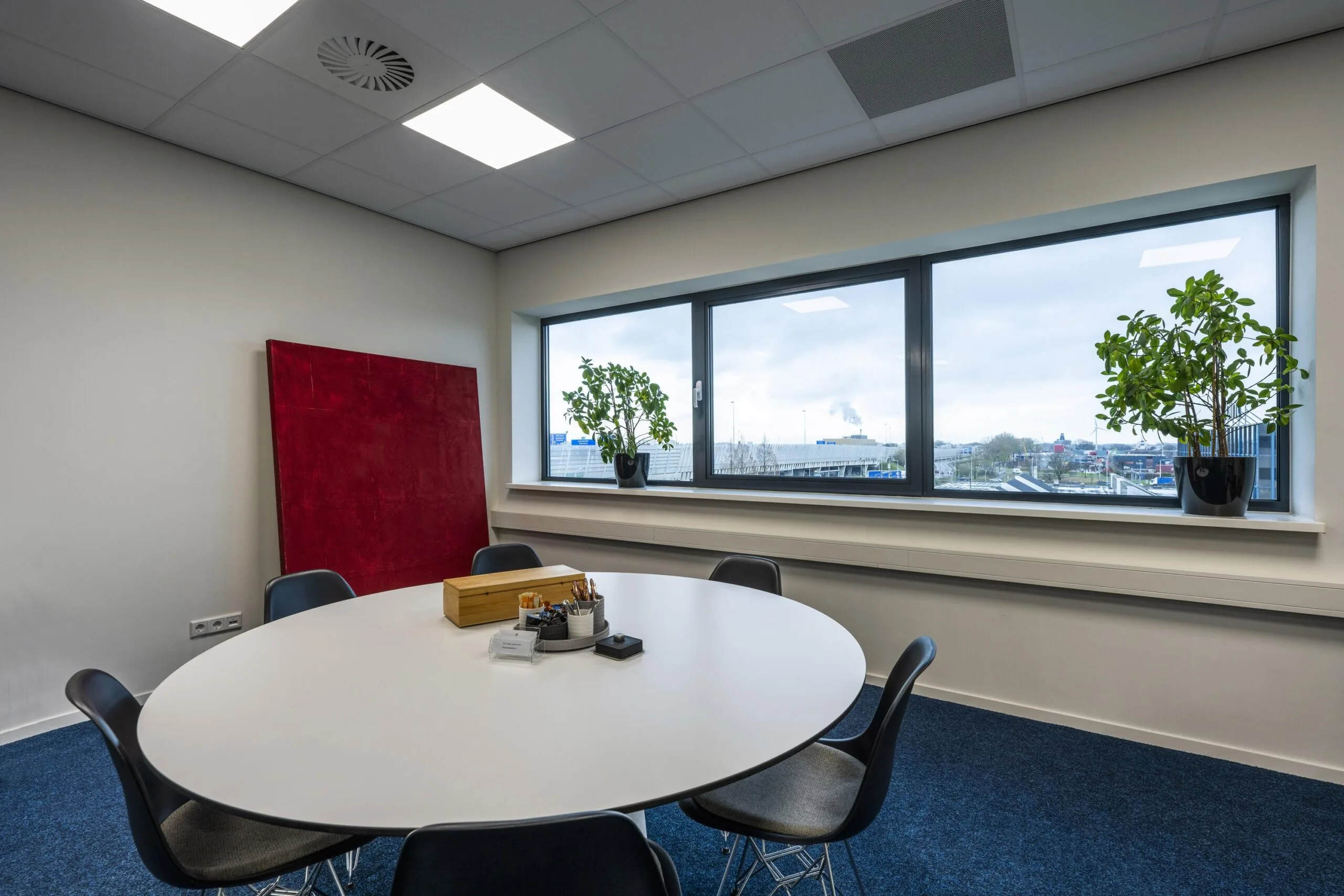 Modern meeting room on Dynamostraat with a round table, black chairs, large window, and two potted plants.
