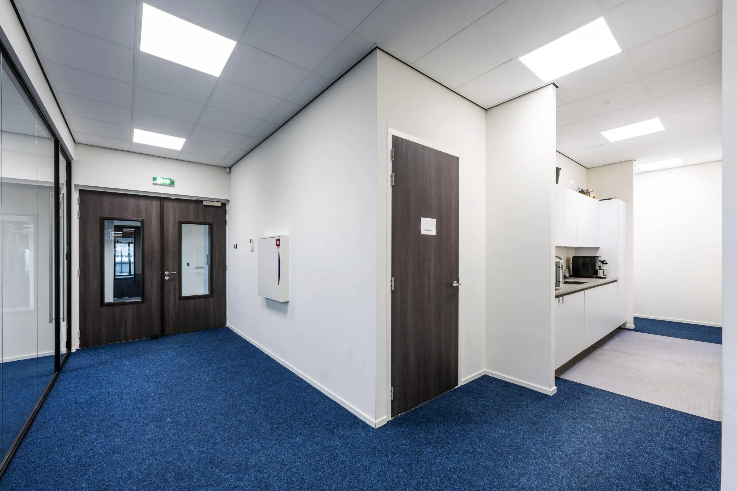 Modern office hallway at Dynamostraat with blue carpet, dark wooden doors, and a kitchenette area.