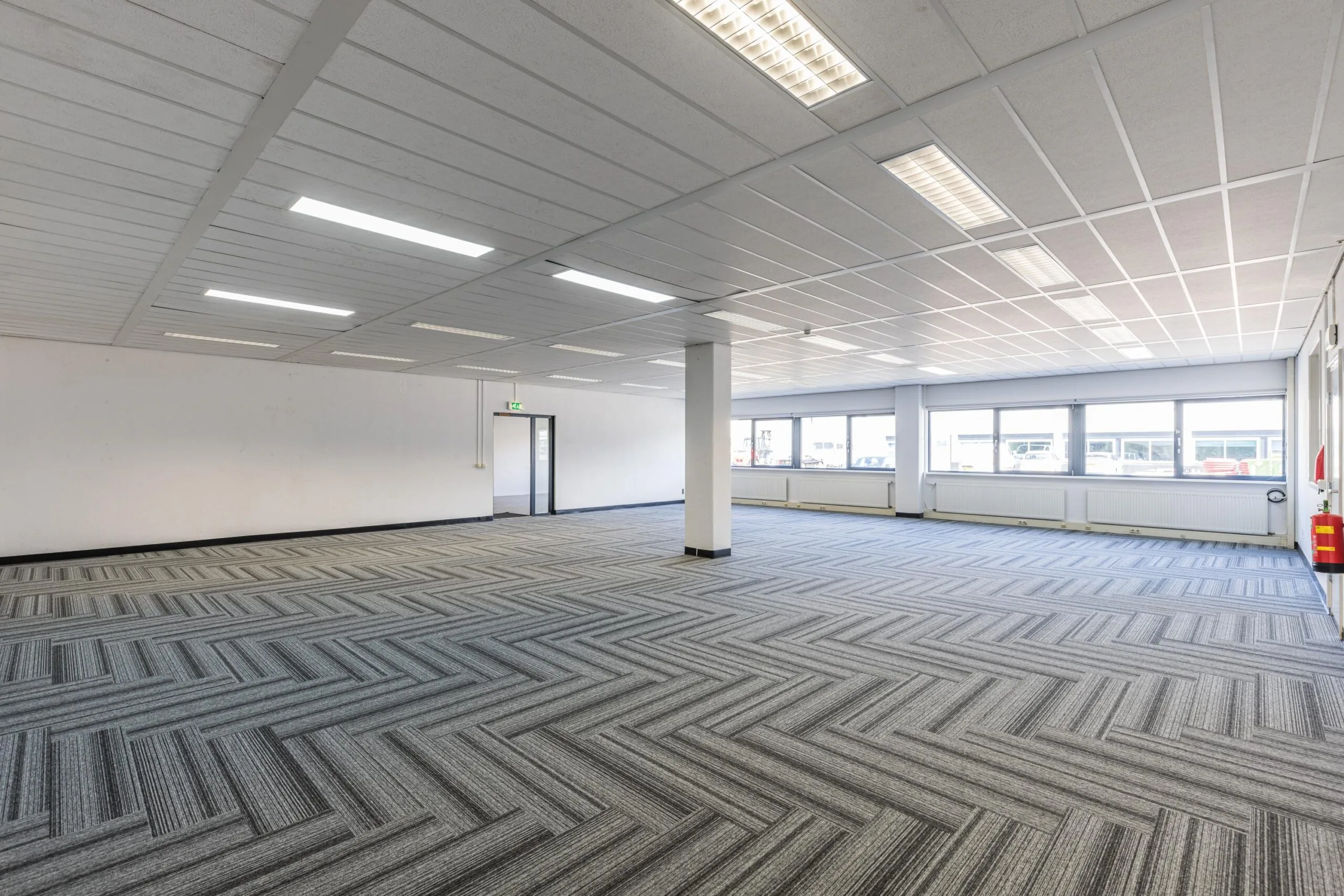Spacious, empty office space on Zonnebaan with large windows, patterned carpet tiles, and fluorescent ceiling lights.