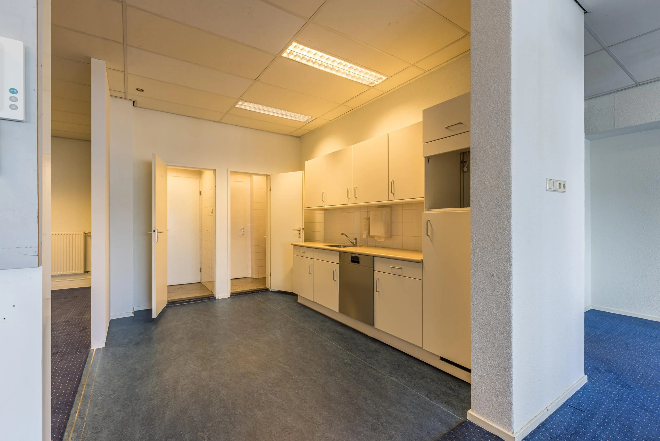 Small office kitchen area with white cabinets, a dishwasher, and three open doors leading to adjacent rooms.