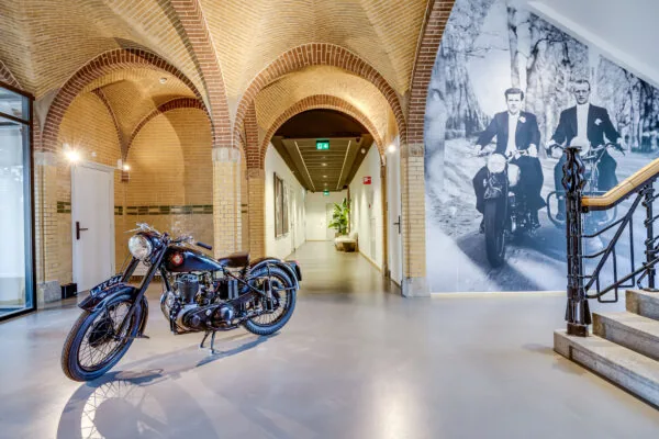 Vintage motorcycle displayed in a hallway with arched brick ceilings and a large black-and-white photo mural of two men riding motorcycles on the wall.