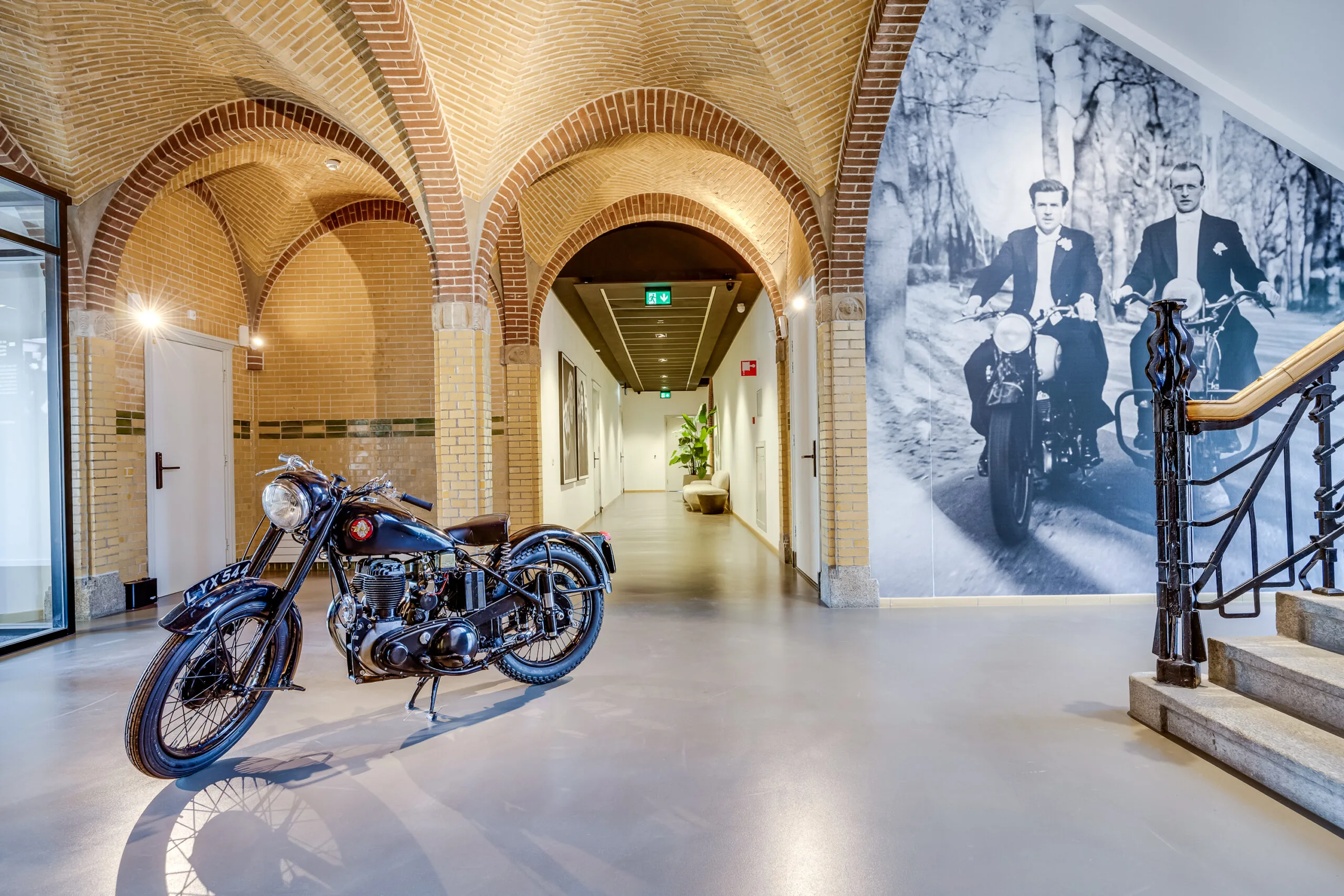 Vintage motorcycle displayed in a hallway with arched brick ceilings and a large black-and-white photo mural of two men riding motorcycles on the wall.