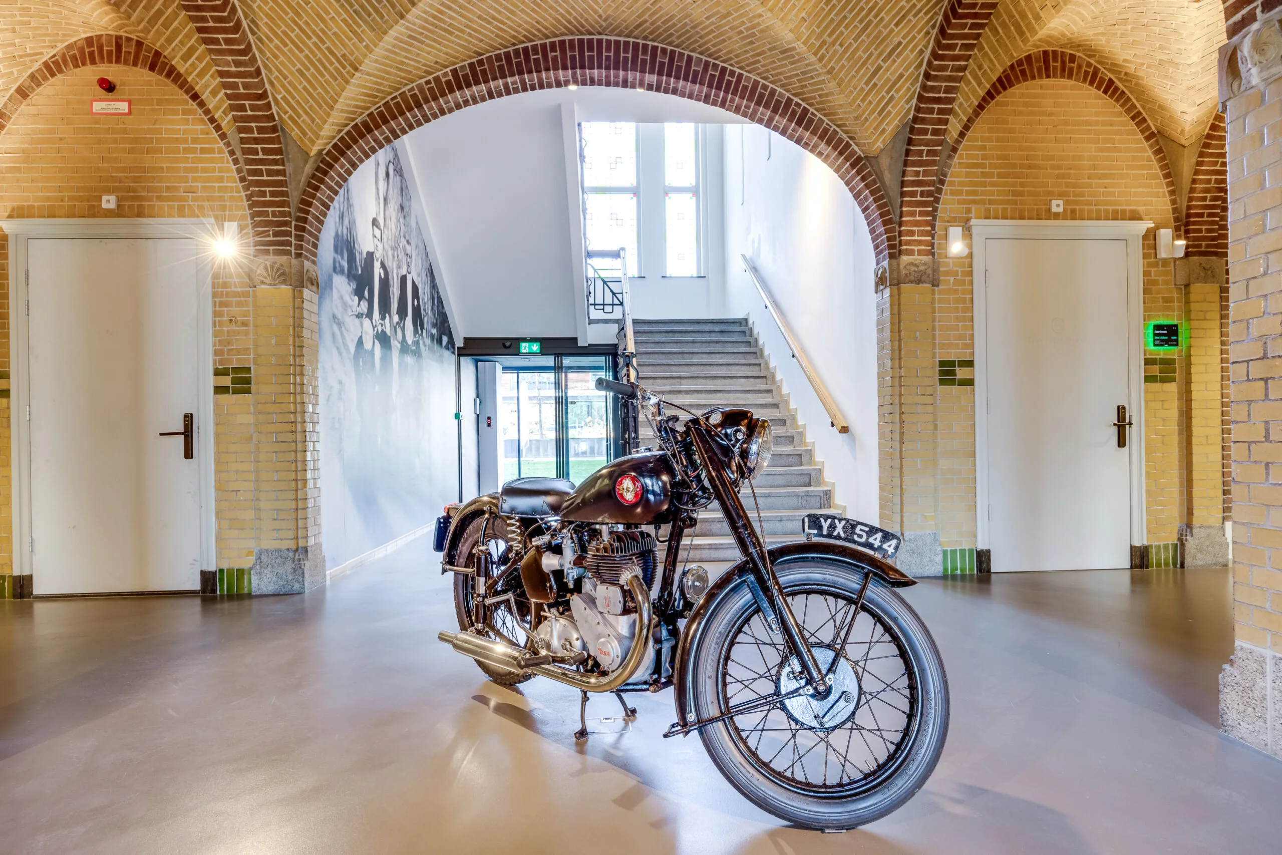 Classic black motorcycle displayed in a bright hallway with arched brick ceilings and a staircase in the background.