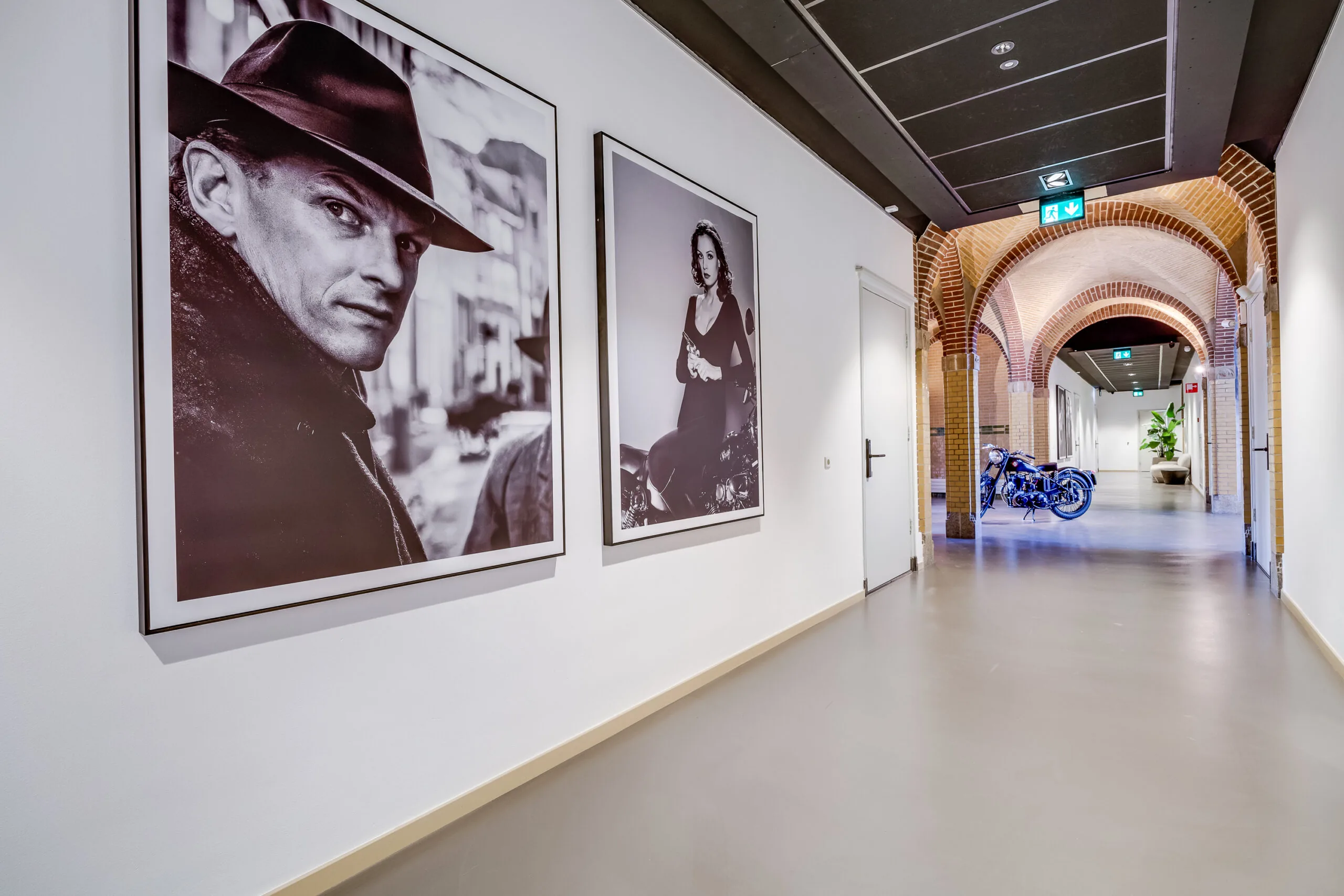 Hallway with large black-and-white portraits on the wall, leading to an arched corridor with a motorcycle and a potted plant.