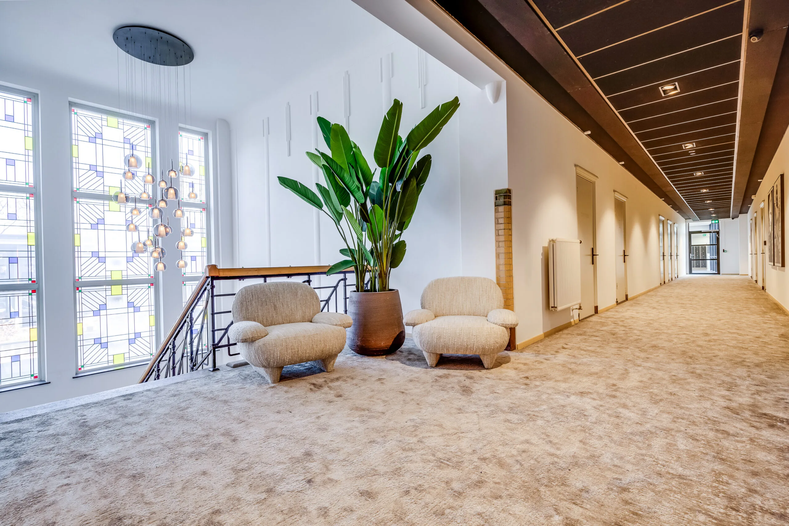 Modern hallway at Lage Naarderweg with two beige lounge chairs, a large potted plant, and colorful stained glass windows.
