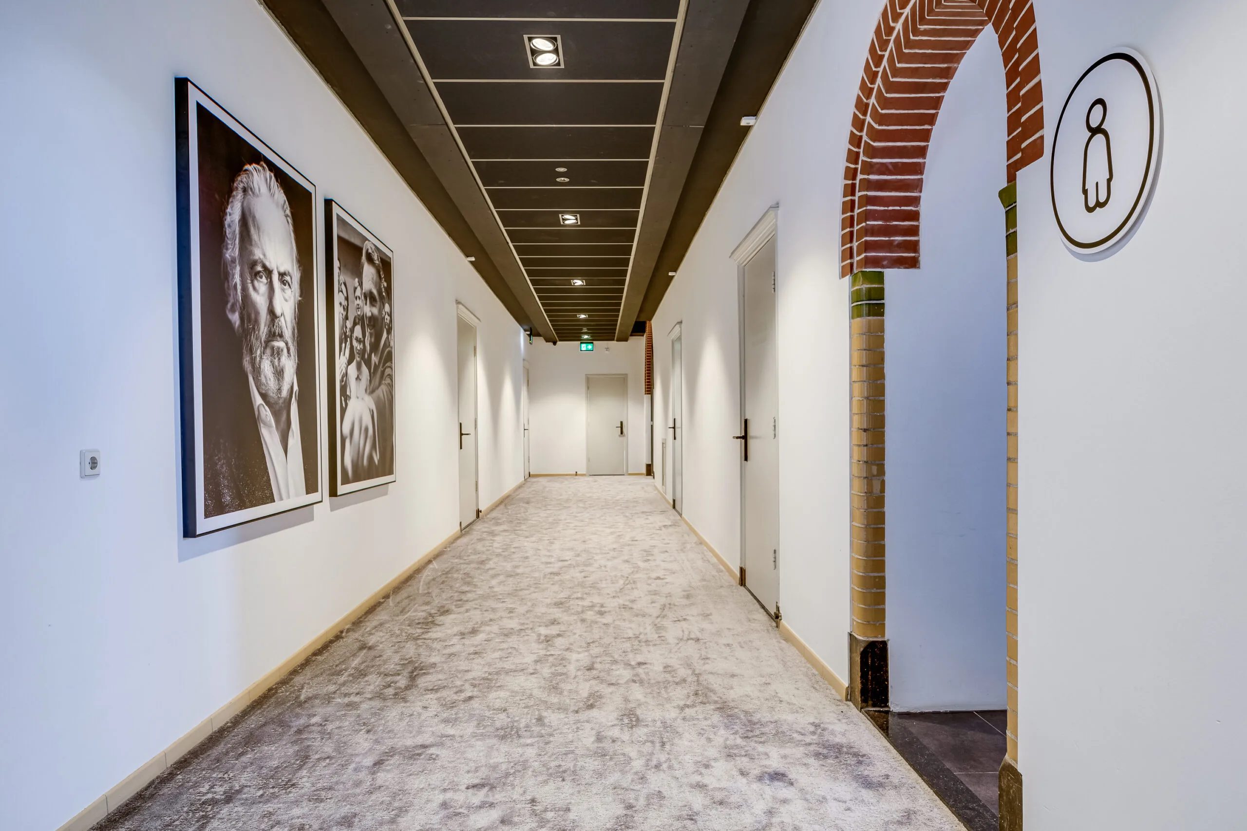 Modern hallway with carpeted floor, black-and-white portraits on the wall, and a women's restroom entrance with a brick arch.