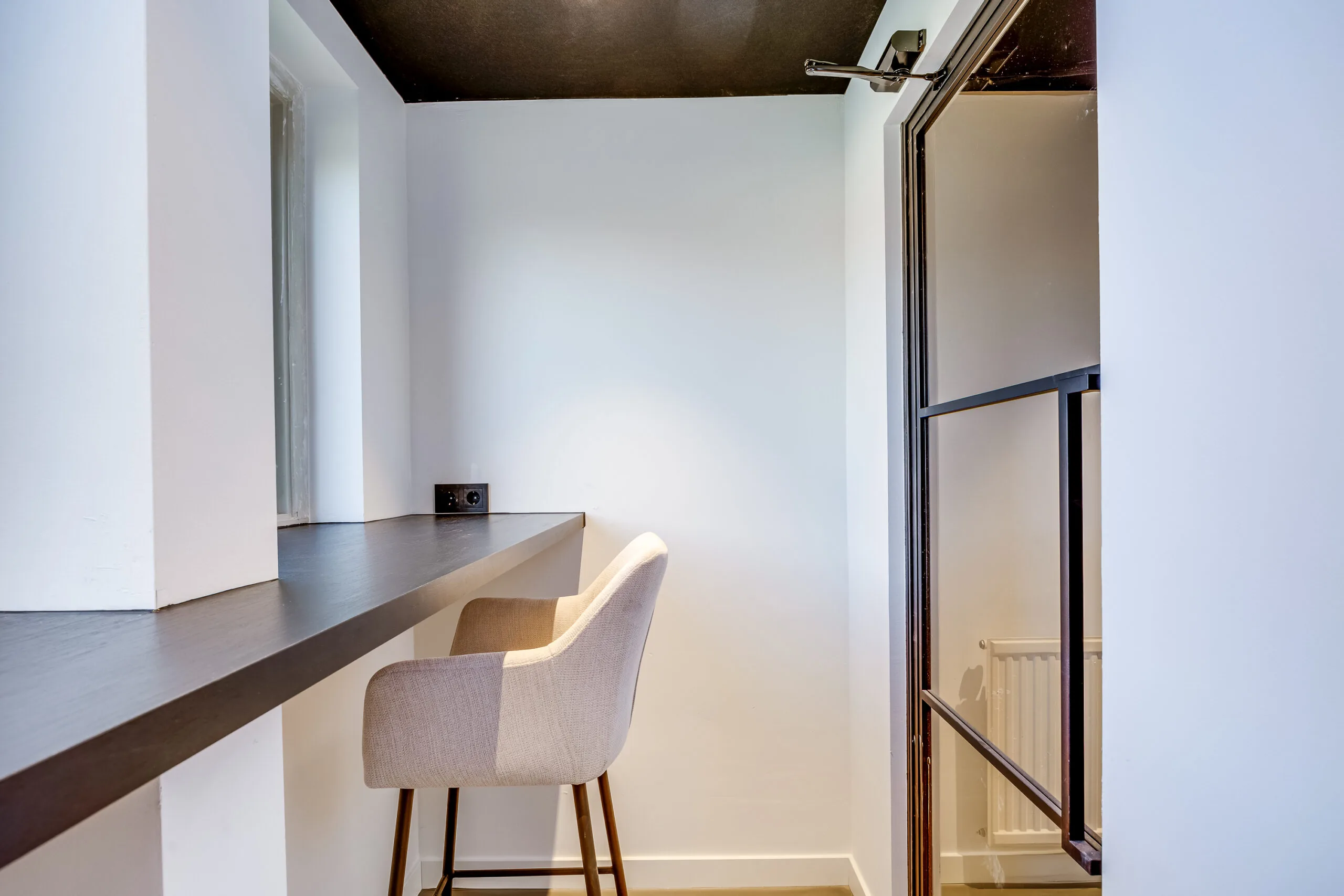Minimalist workspace with a built-in dark wooden desk, a beige chair, and a black-framed glass door at Lage Naarderweg.