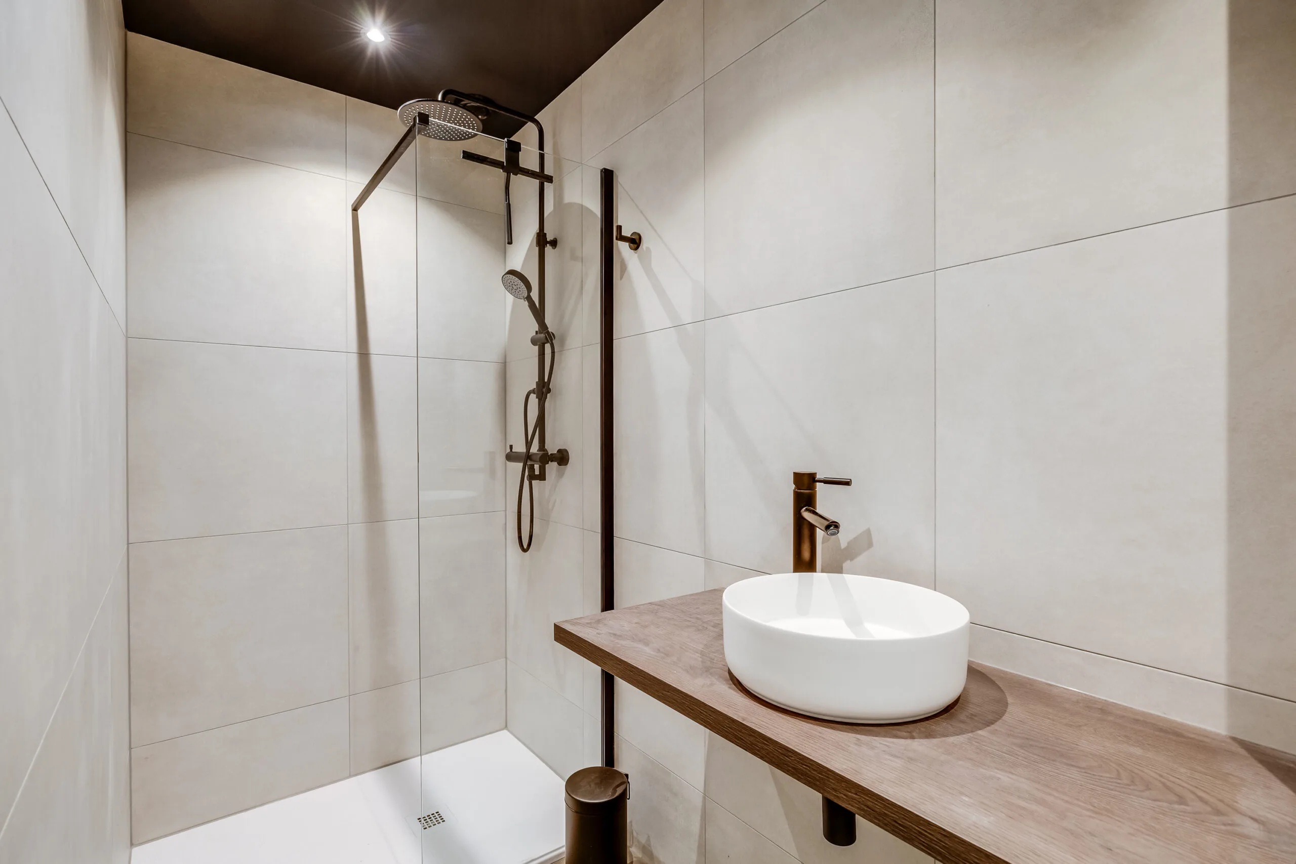 Modern walk-in shower with beige tiled walls, a rainfall showerhead, and a minimalist wooden vanity with a round white basin.