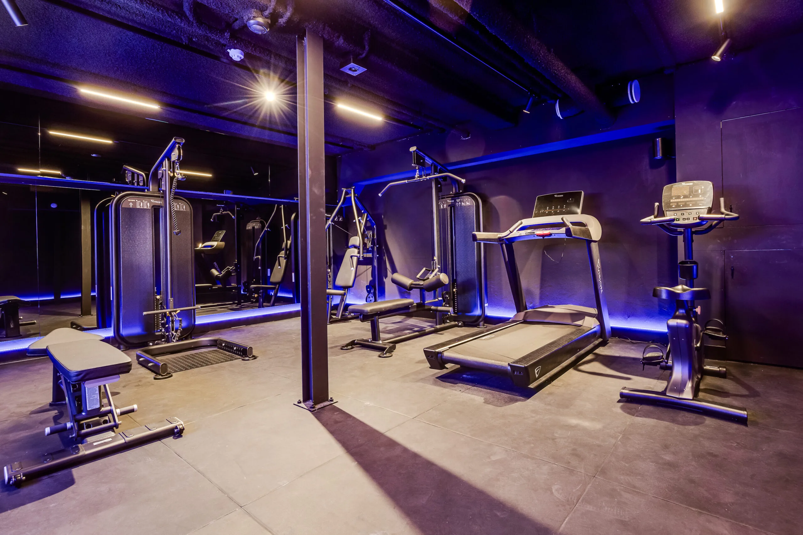 Modern gym with various fitness machines, including a treadmill, exercise bike, and weight equipment, in a sleek, dark interior on Lage Naarderweg.