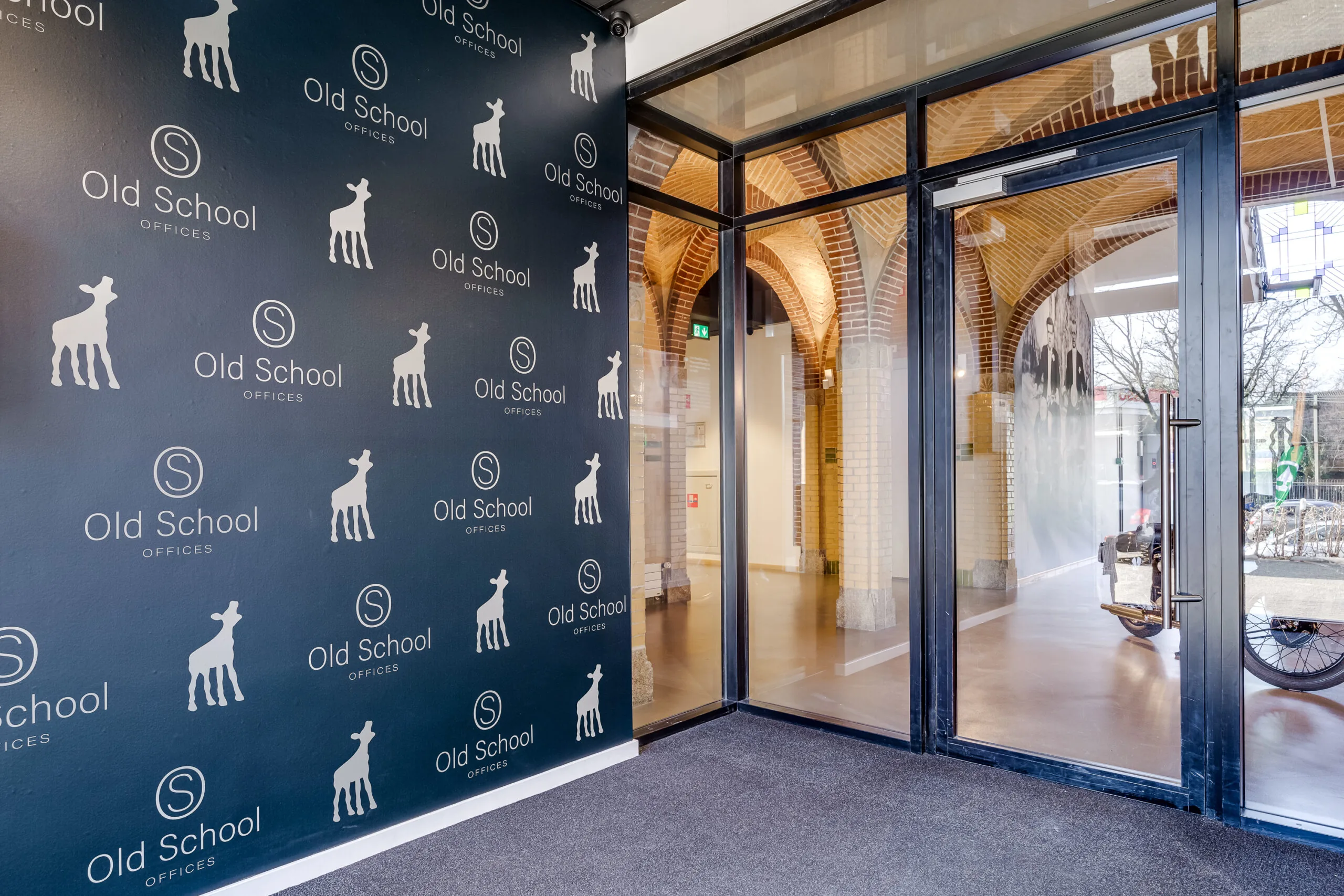 Entrance area of Old School Offices at Lage Naarderweg, featuring a logo wall and glass doors leading to a brick-arched hallway.