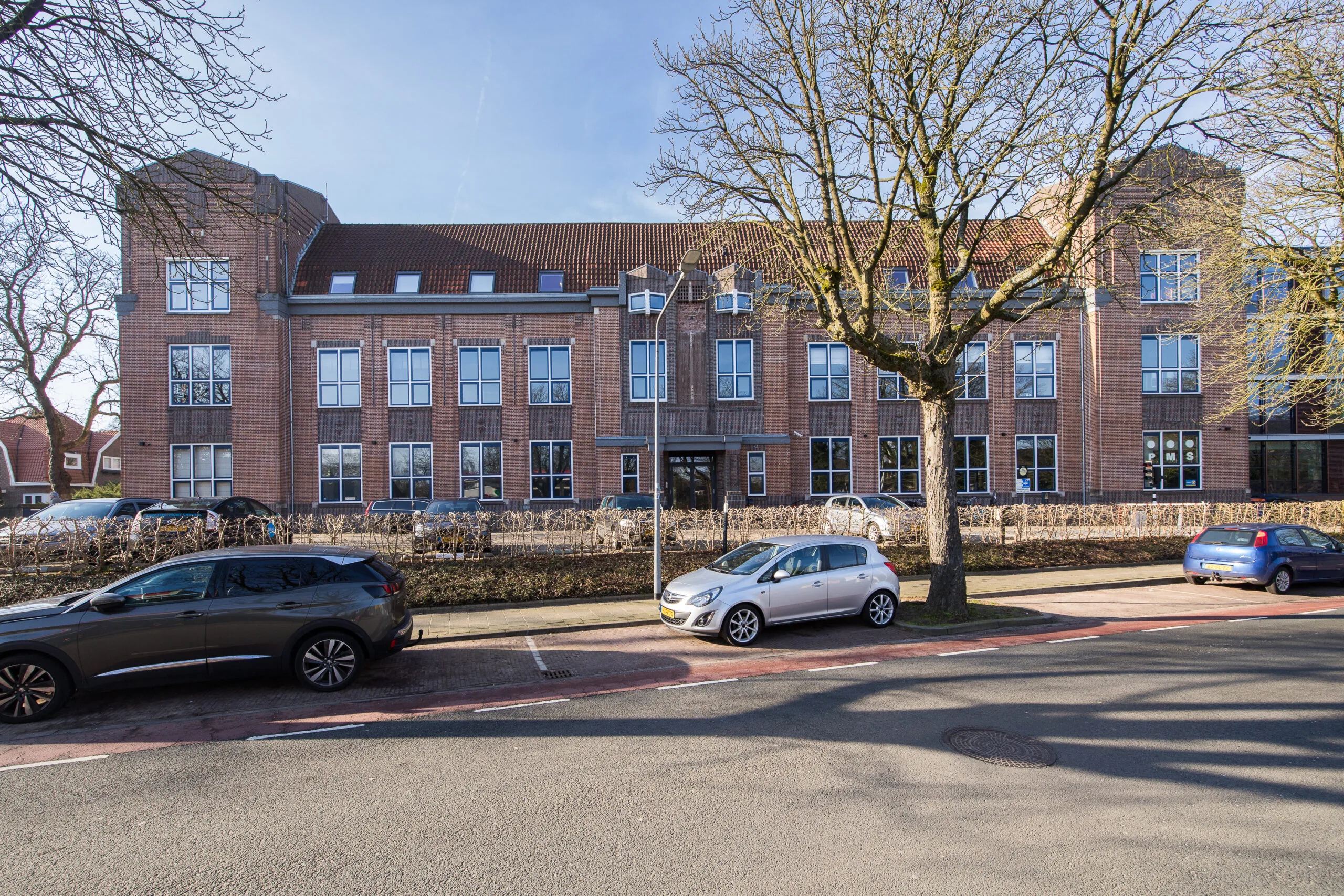 Front view of a large brick building on Lage Naarderweg with parked cars and leafless trees.