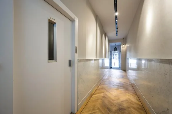 Verlichte gang in een pand aan de Nieuwe Spiegelstraat met een witte liftdeur en visgraat houten vloer.