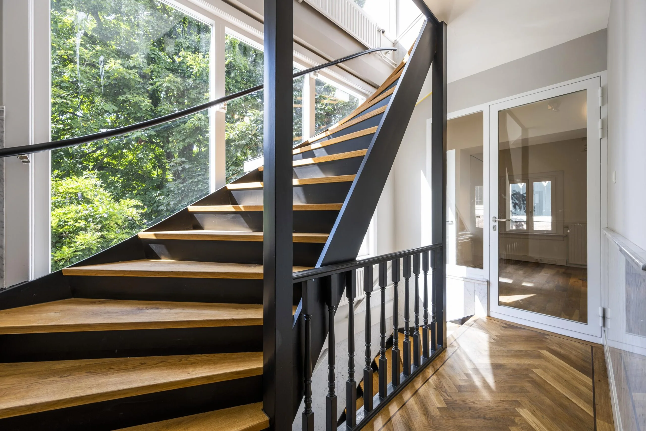 Moderne houten trap met zwarte leuningen en grote ramen in een licht trappenhuis aan de Nieuwe Spiegelstraat.