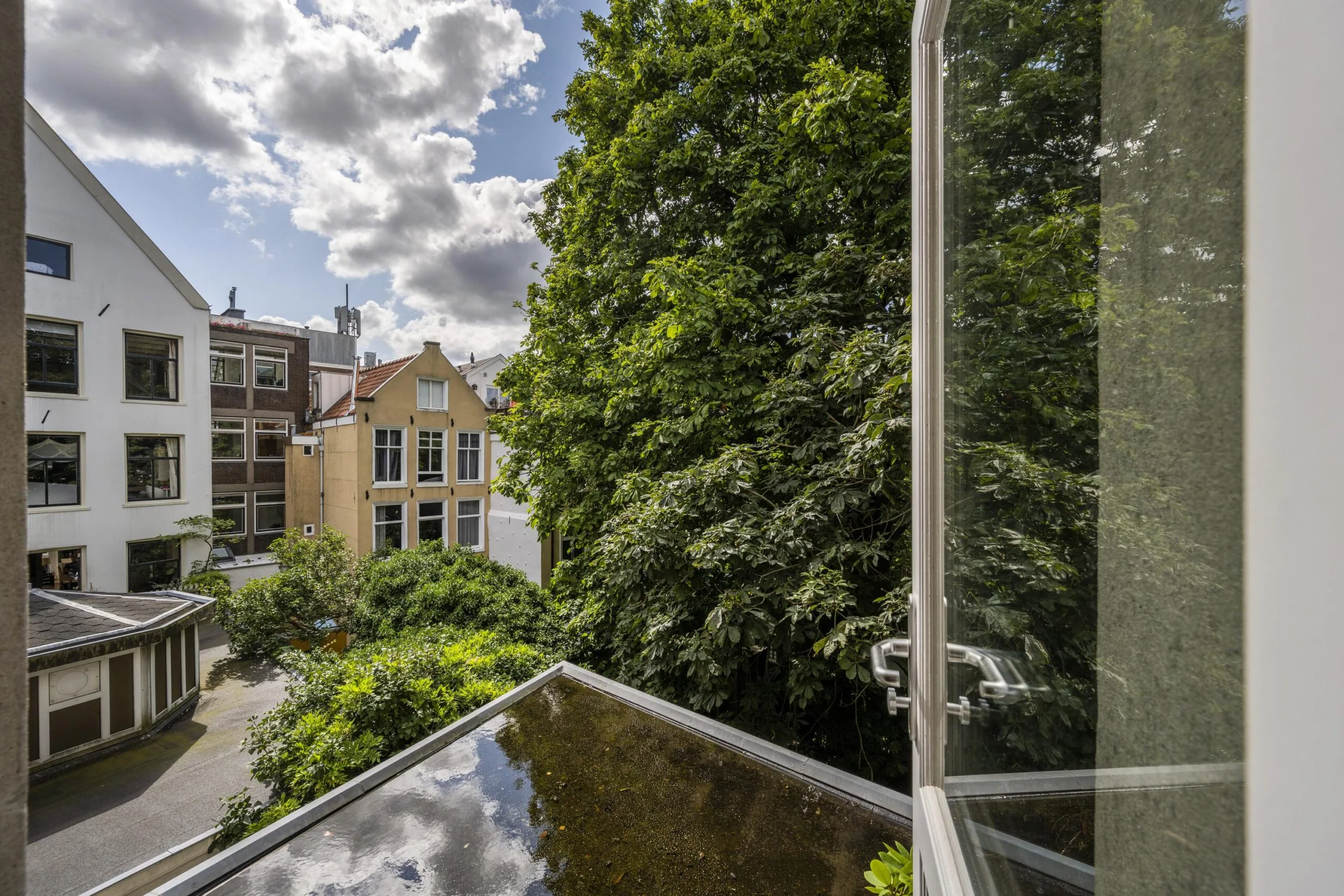 Uitzicht vanuit een open raam op de Nieuwe Spiegelstraat met bomen, stadspanden en een dakterras.