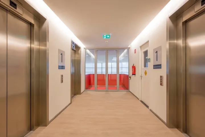 Hal van een modern kantoorgebouw op de eerste verdieping met twee liften en een glazen deur naar de Stationsstraat.