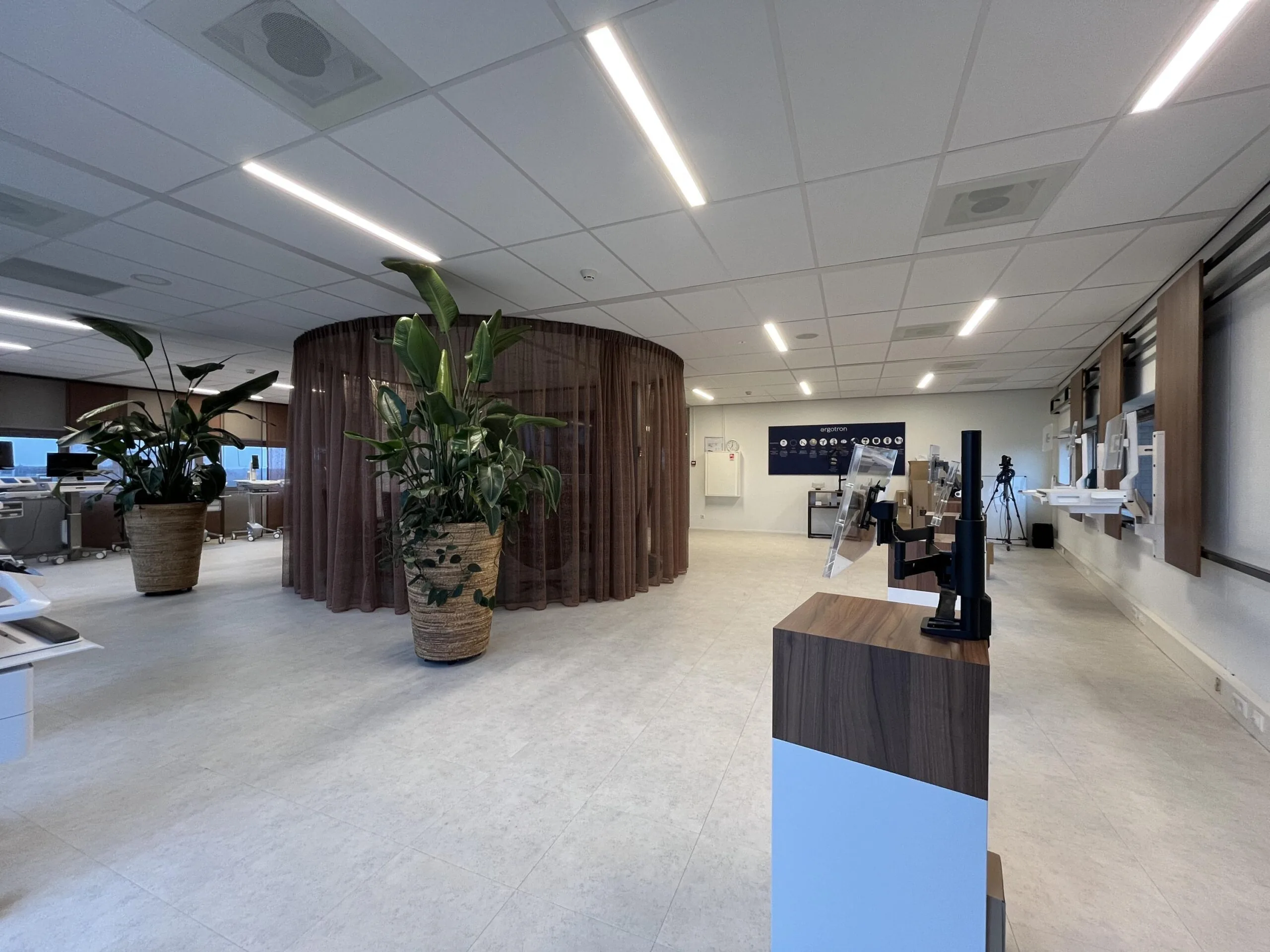 Moderne, ruim opgezette kantoor- of zorgomgeving aan de Stationsstraat met bureaus, grote planten en een ronde, halftransparante afscheiding.