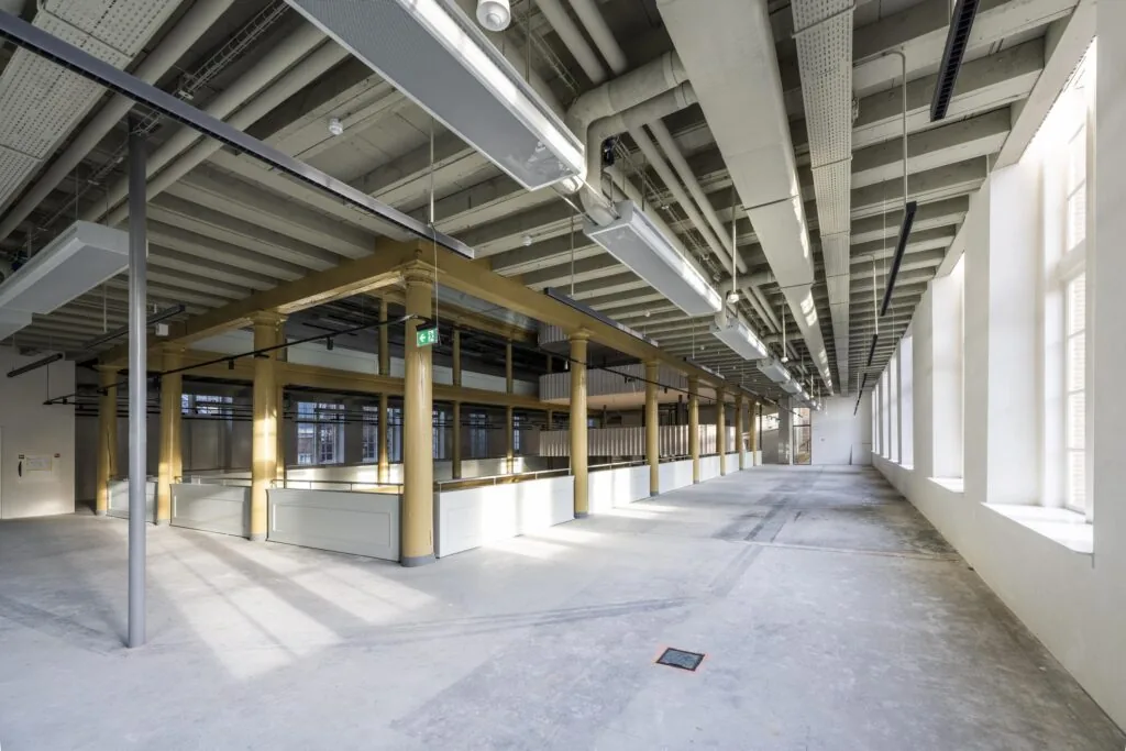 Lichte, industriële ruimte in het gebouw aan de Kloveniersburgwal met betonnen vloer, witte muren en zichtbare leidingen aan het plafond.