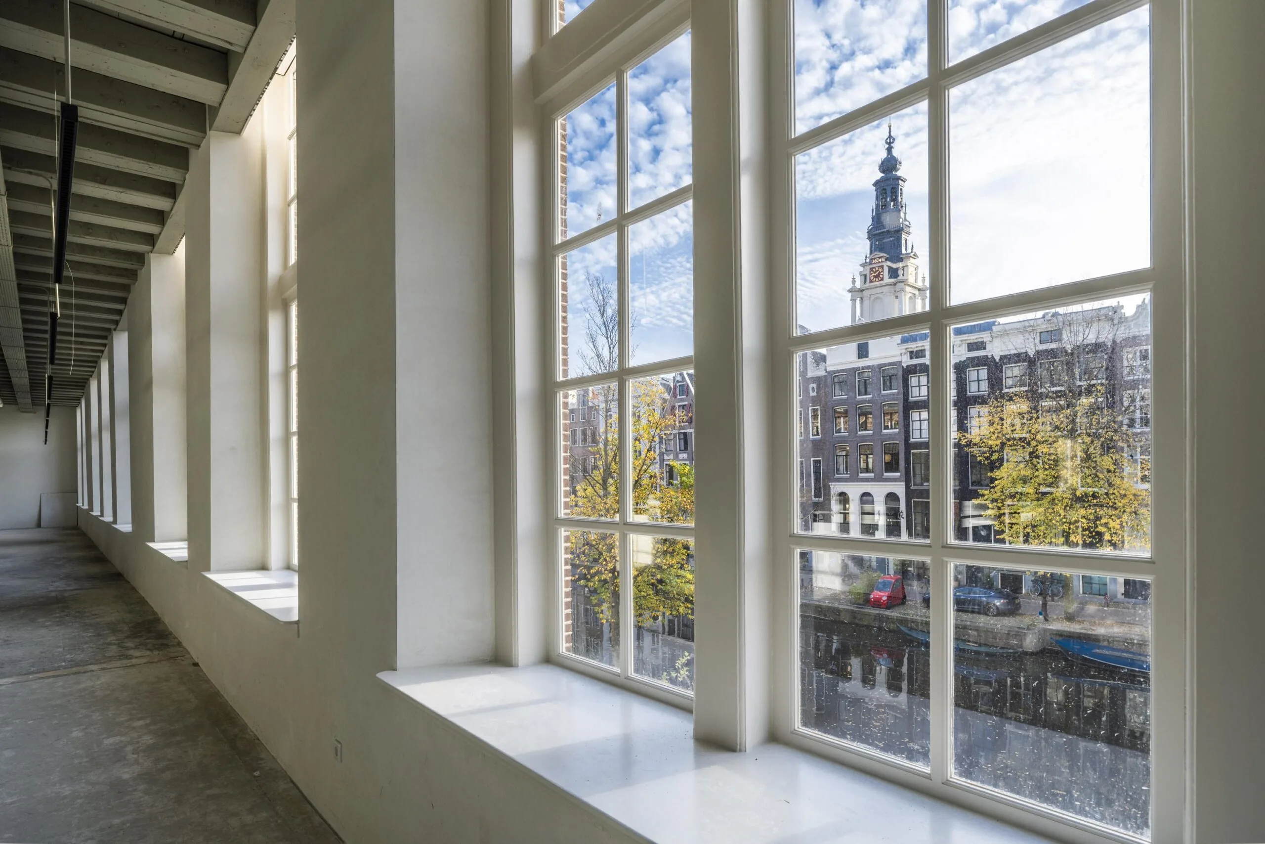 Uitzicht vanuit een leeg interieur aan de Kloveniersburgwal op Amsterdamse grachtenpanden en de Zuiderkerkstoren.