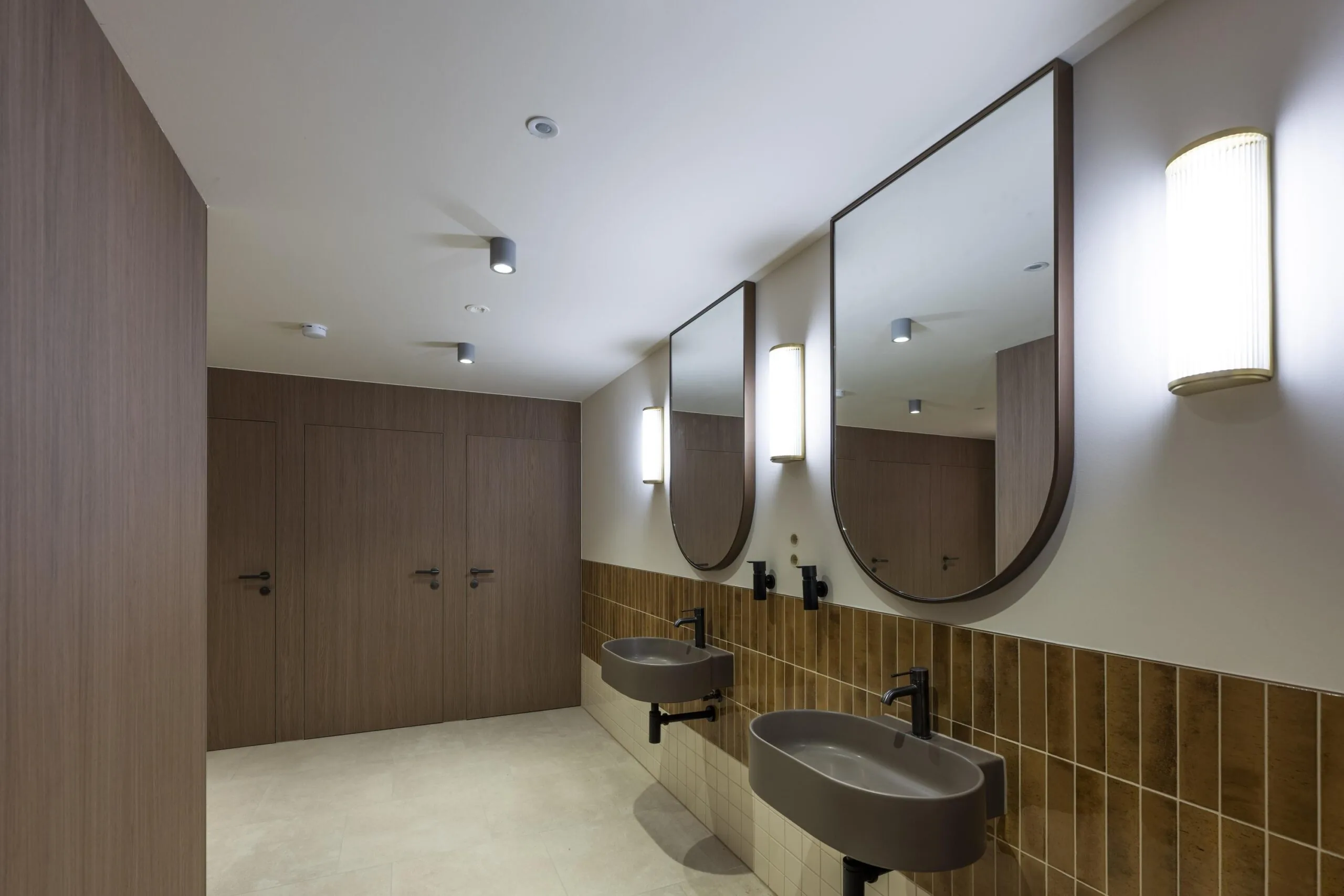 Moderne openbare toiletruimte met houten deuren, twee wastafels en grote spiegels aan de muur.