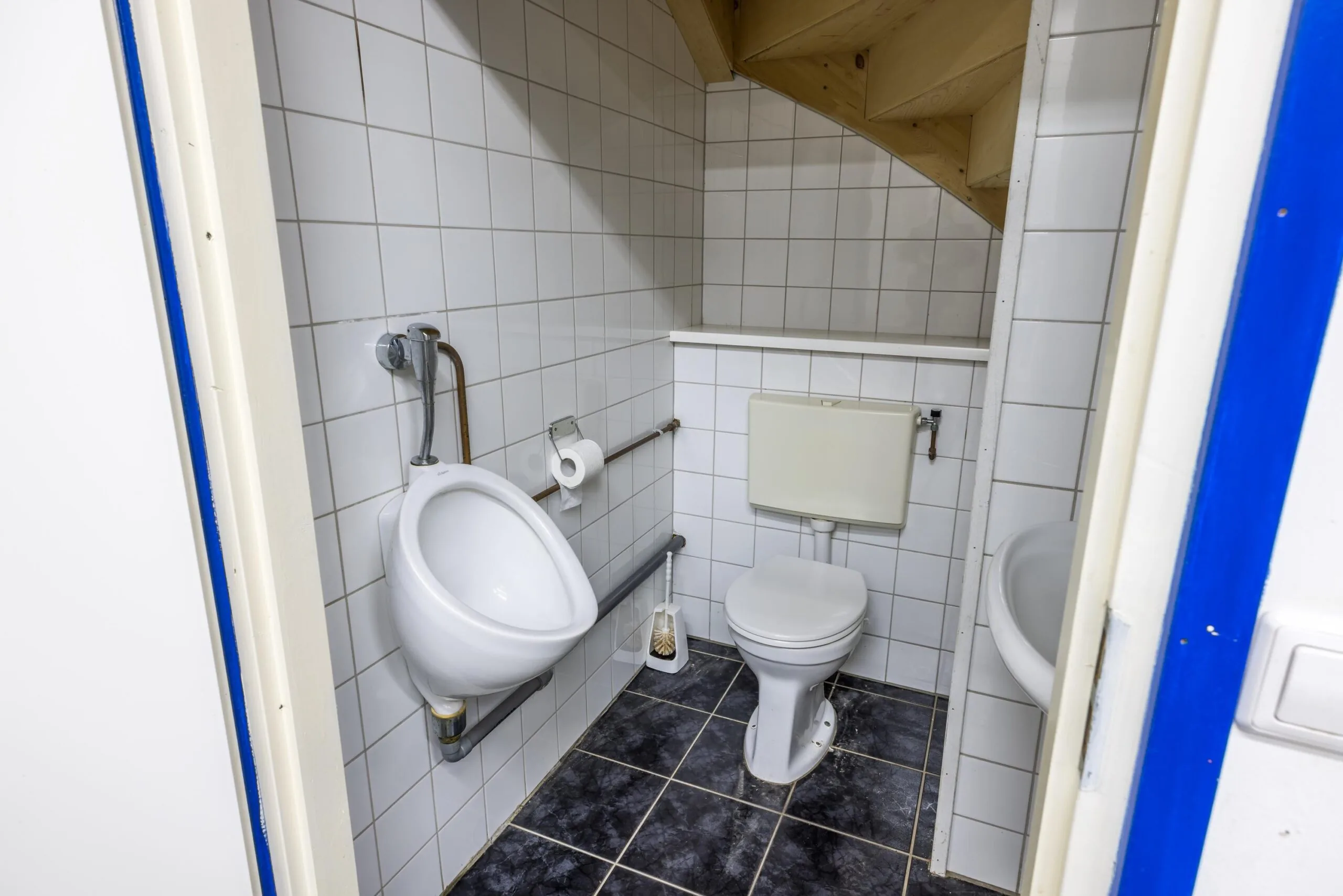 Kleine betegelde toiletruimte aan de Tennesseedreef met een toilet, urinoir en wastafel onder een houten trap.