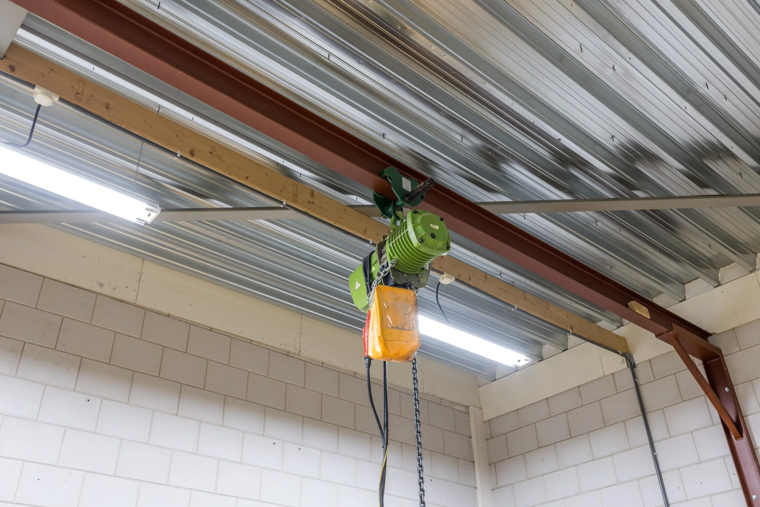 Groene takel met ketting bevestigd aan een rode stalen balk onder het plafond van een loods aan de Tennesseedreef.
