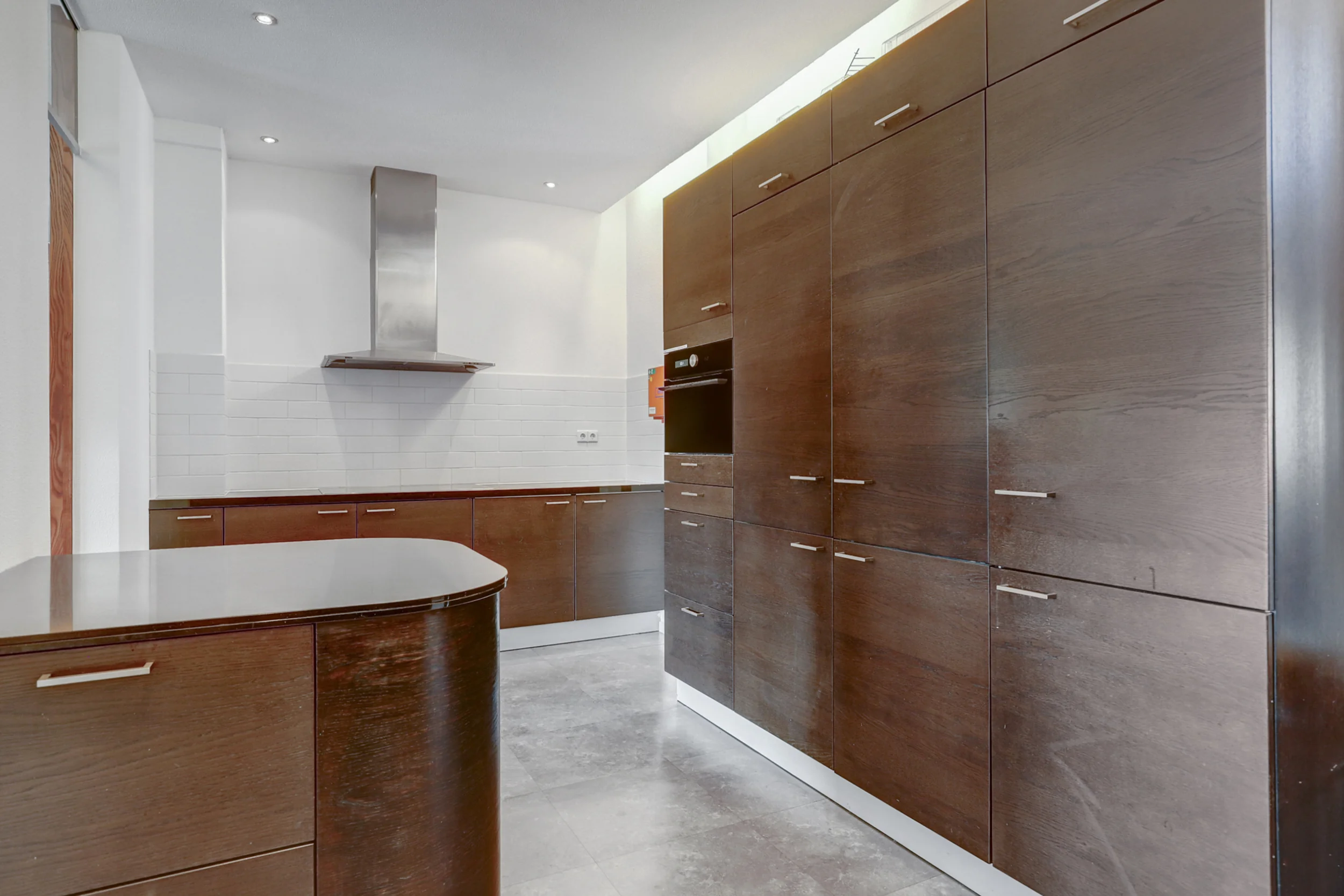 Moderne keuken met donkerhouten kastdeuren, een ingebouwde oven en een roestvrijstalen afzuigkap tegen een witte tegelwand.