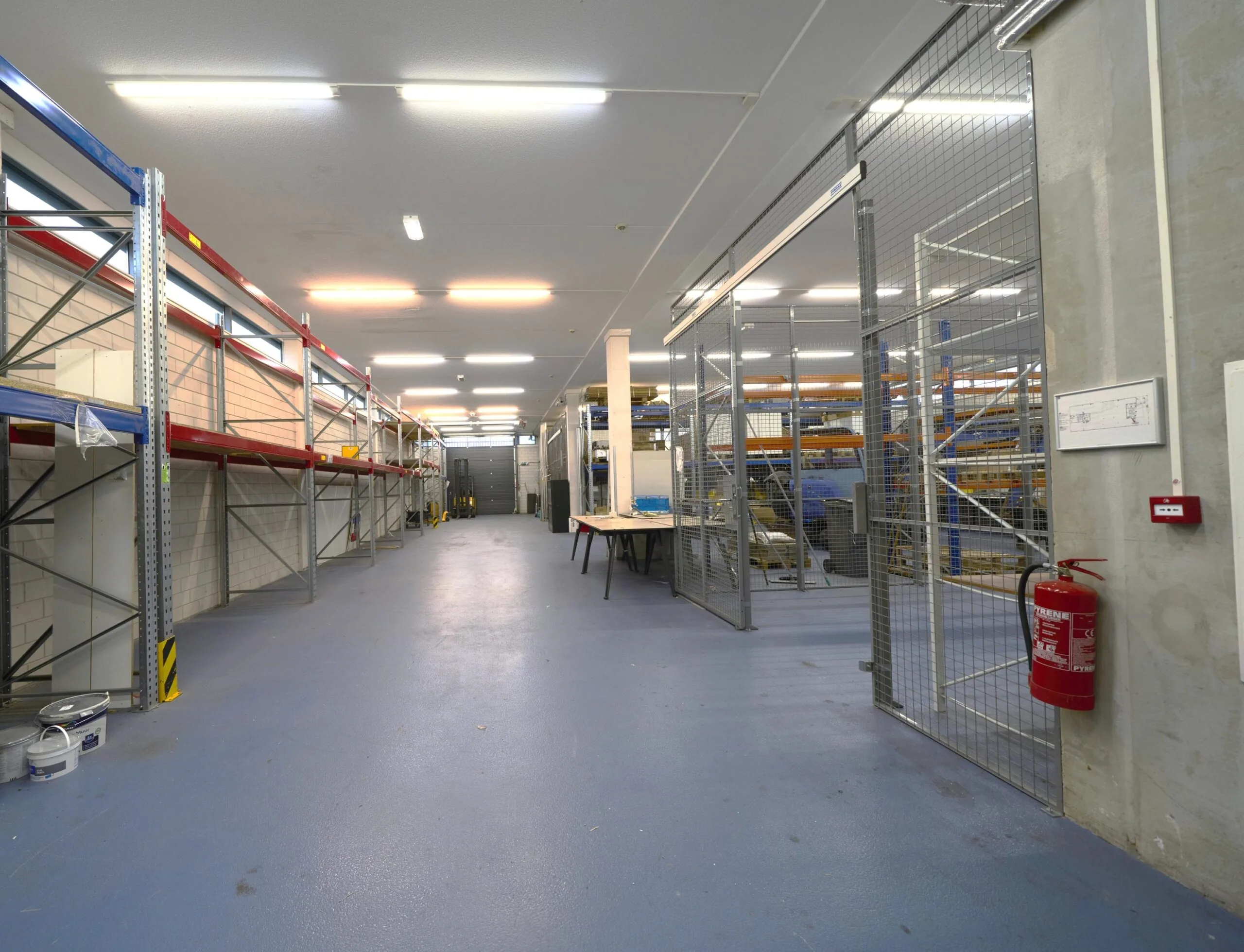 Magazijnruimte met lege metalen stellingen, een afgesloten roosterafdeling en een brandblusser aan de muur.
