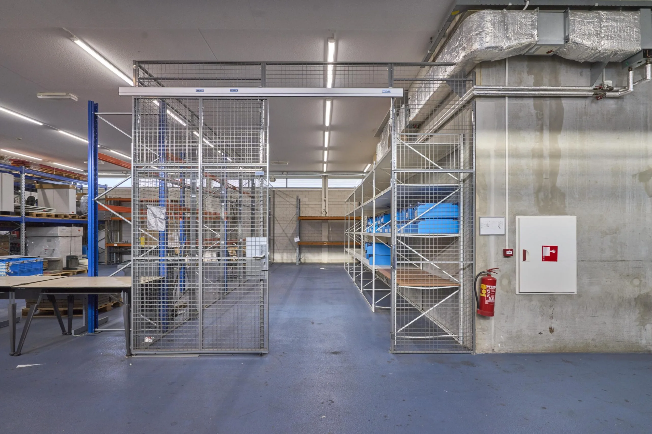 Magazijnruimte met metalen stellingen, blauwe opbergboxen en een brandblusser aan de betonnen muur.