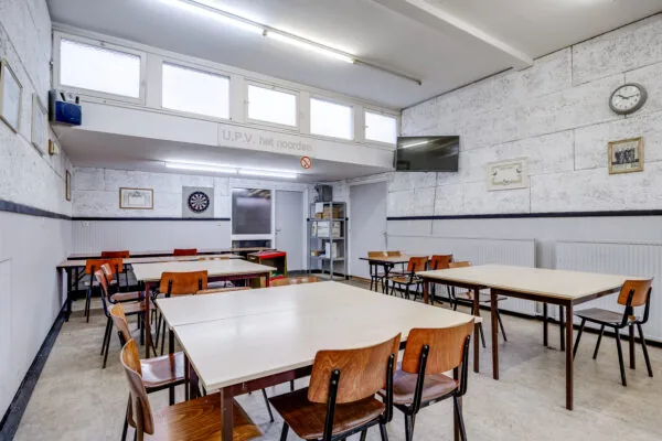 Gemeentelijke vergaderruimte of kantine met tafels, stoelen, een dartbord en een televisie, gelegen aan de Balderikstraat.