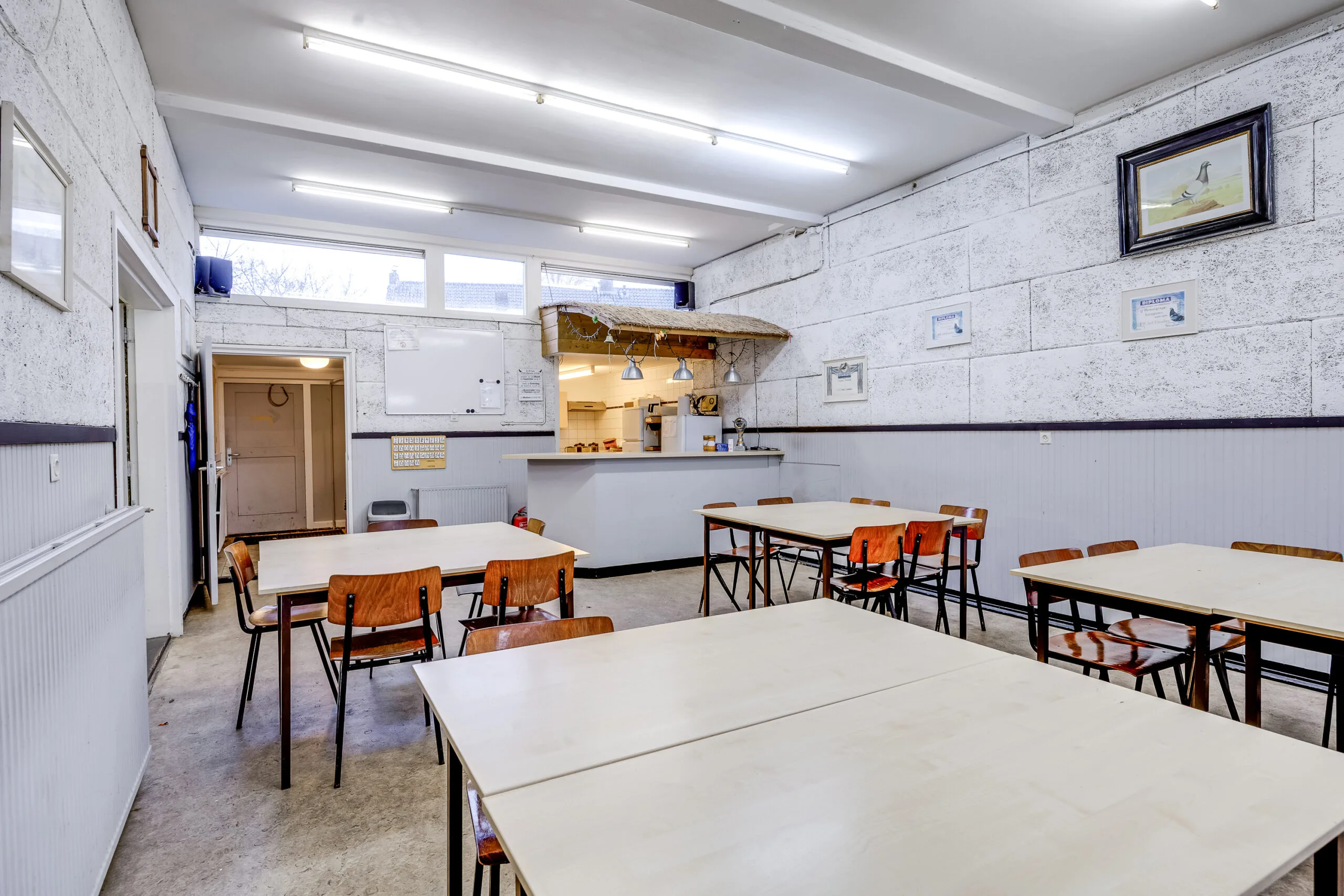 Eenvoudige kantineruimte met houten tafels en stoelen, een bar met rieten dakje en versierde muren in een gebouw aan de Balderikstraat.