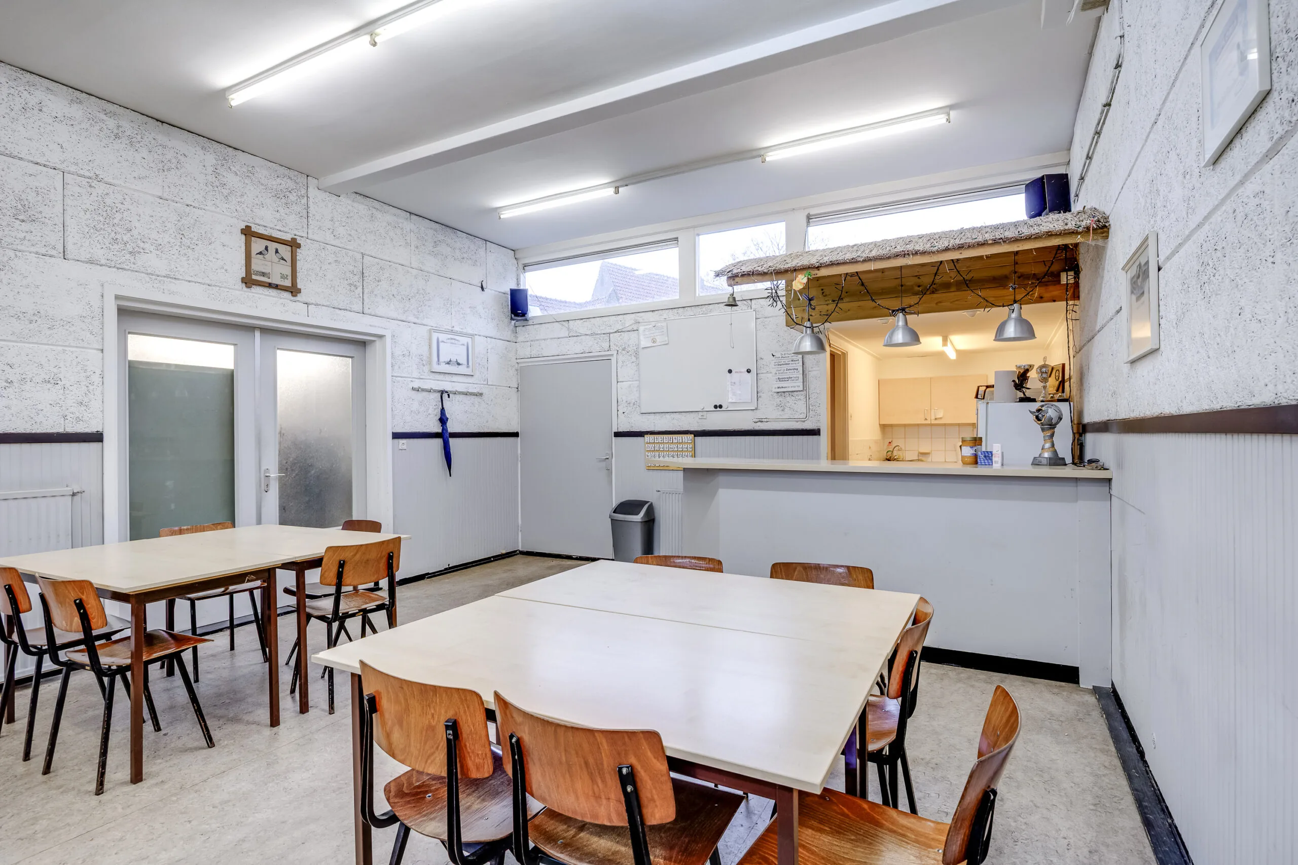 Gemeenschappelijke ruimte met eenvoudige houten tafels en stoelen, een bar met keuken en TL-verlichting aan het plafond in de Balderikstraat.