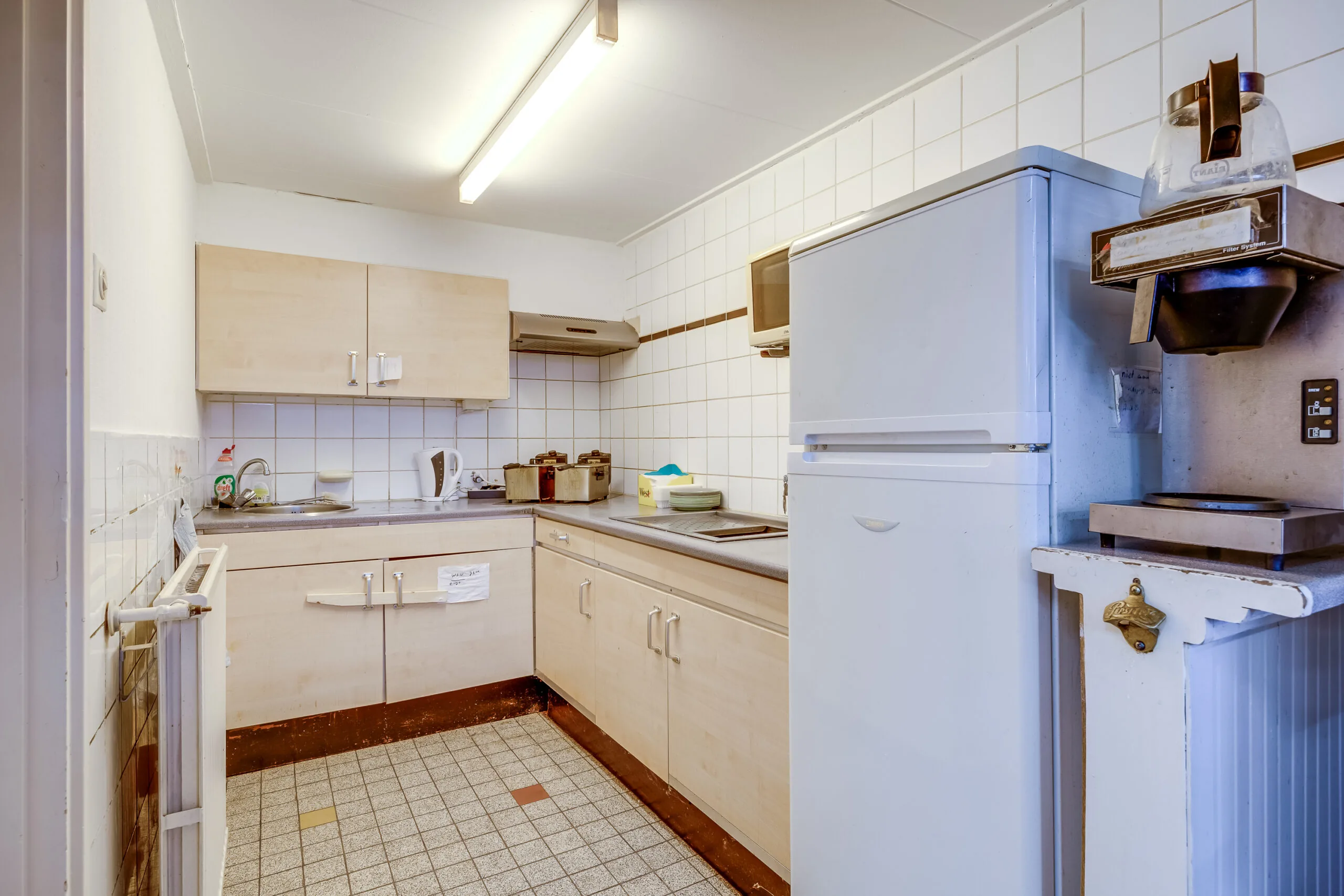 Keuken met licht houten kastjes, wit tegelwerk, een koelkast, koffiezetapparaat en diverse keukenapparatuur in de Balderikstraat.