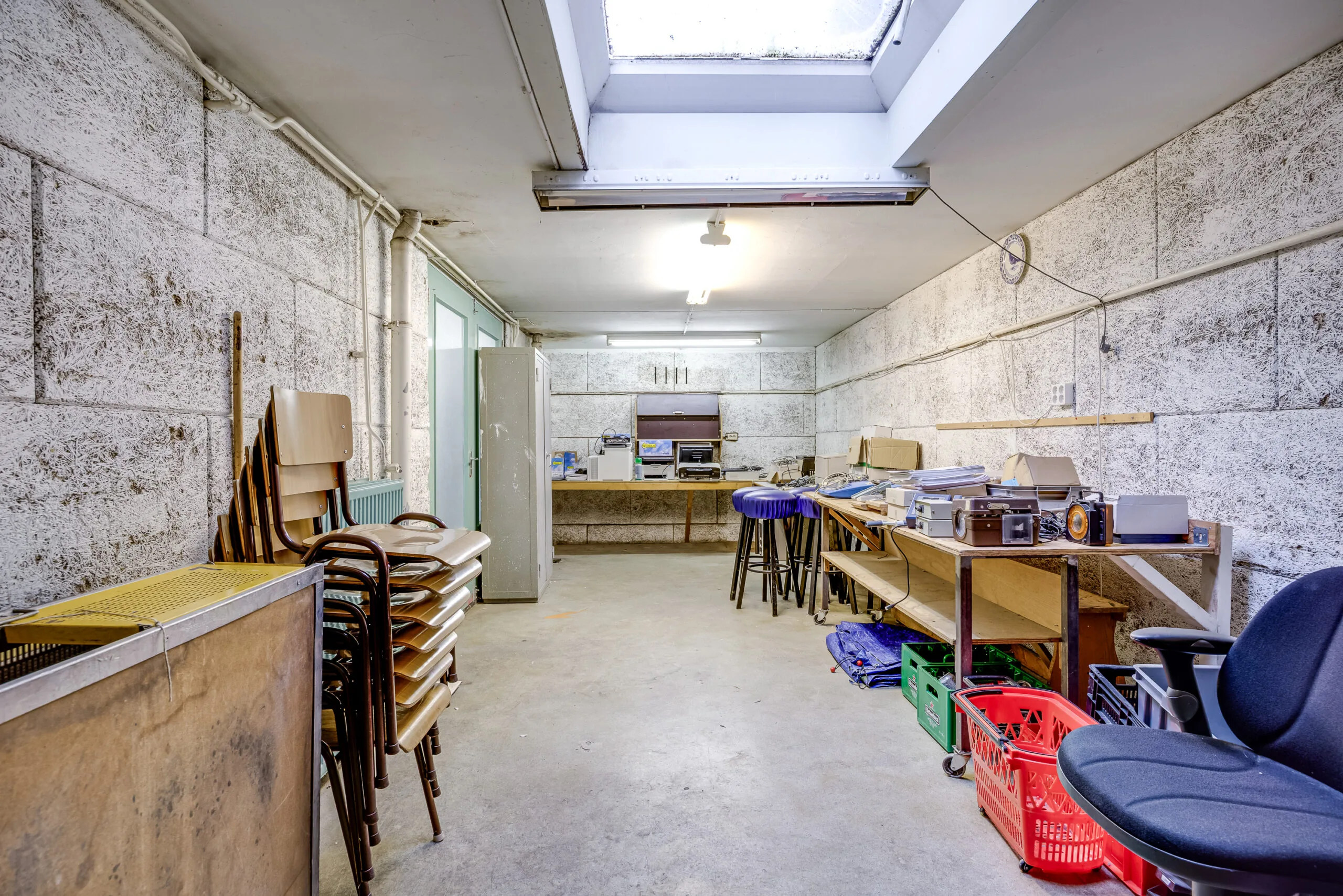 Opslagruimte aan de Balderikstraat met stapelstoelen, werktafels vol apparatuur en een groot dakraam.