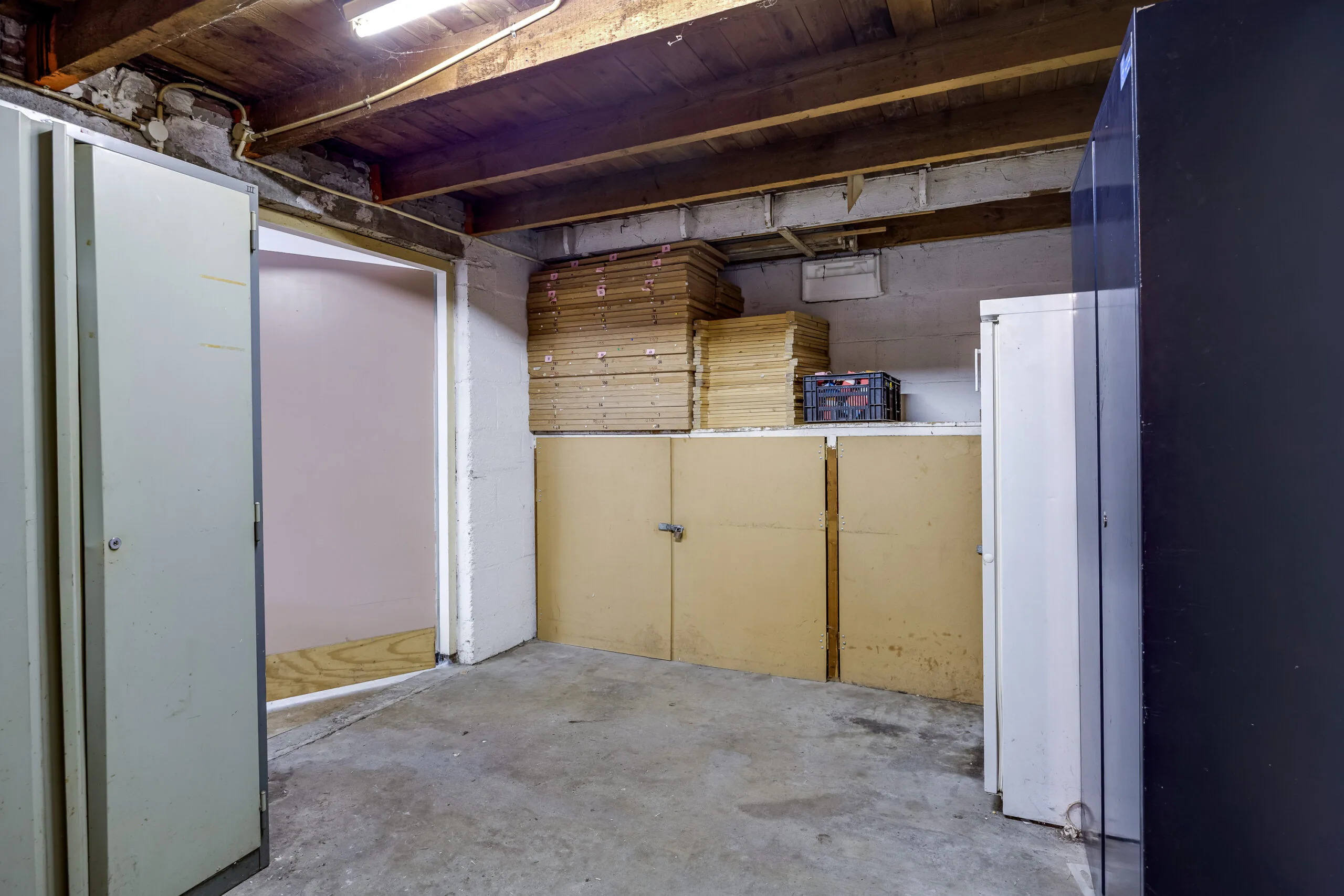 Opslagruimte in een kelder met betonnen vloer, houten plafond, opgestapelde houten planken en diverse kasten.
