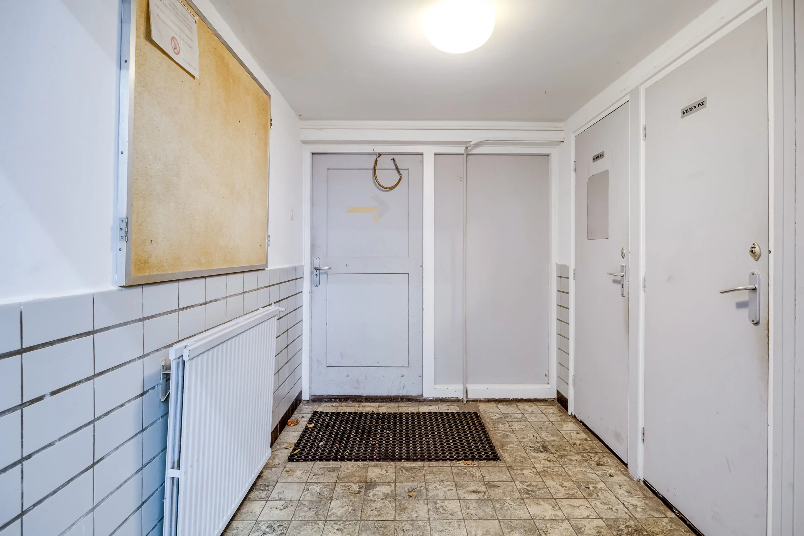 Entreehal met drie witte deuren, een radiator, een prikbord en een vloer met verouderde tegels in een gebouw aan de Balderikstraat.