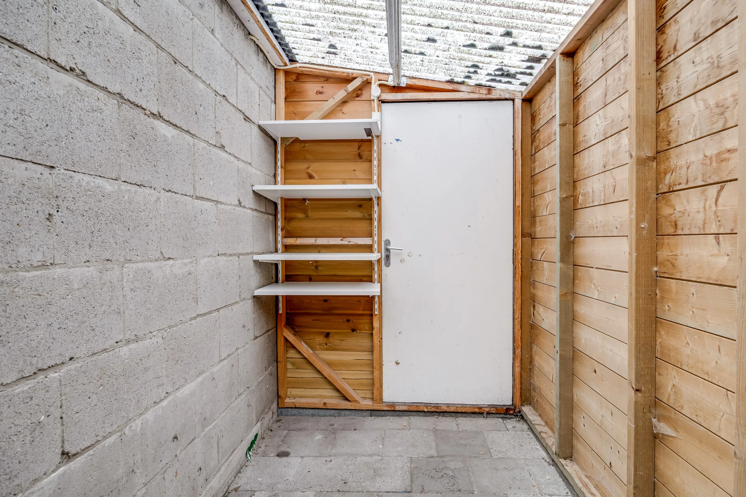Houten berging met betonnen muur, witte deur en hangende witte wandplanken aan de Balderikstraat.