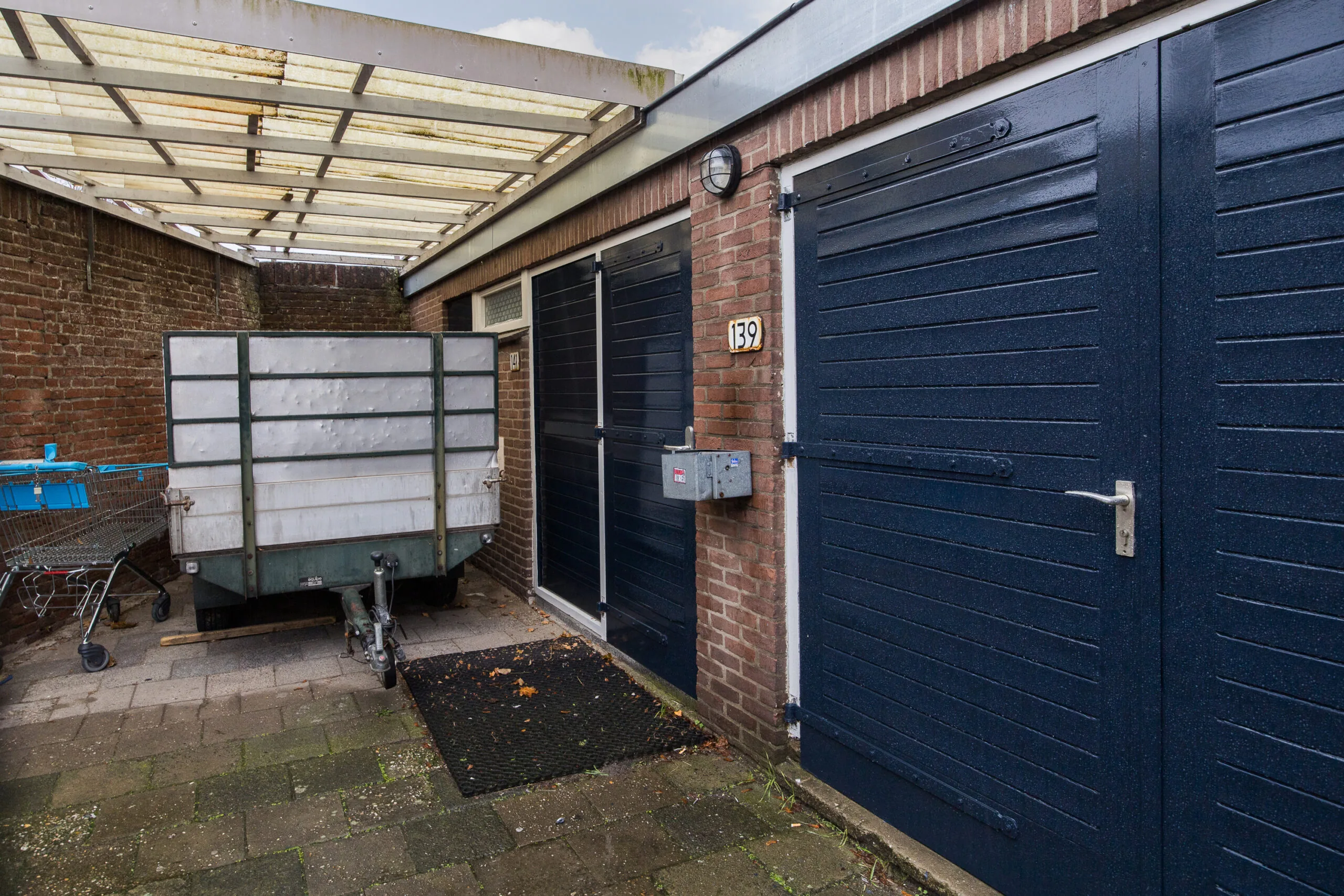 Overdekte achterom met blauwe garagedeuren, een aanhanger en huisnummer 139 aan de Balderikstraat.