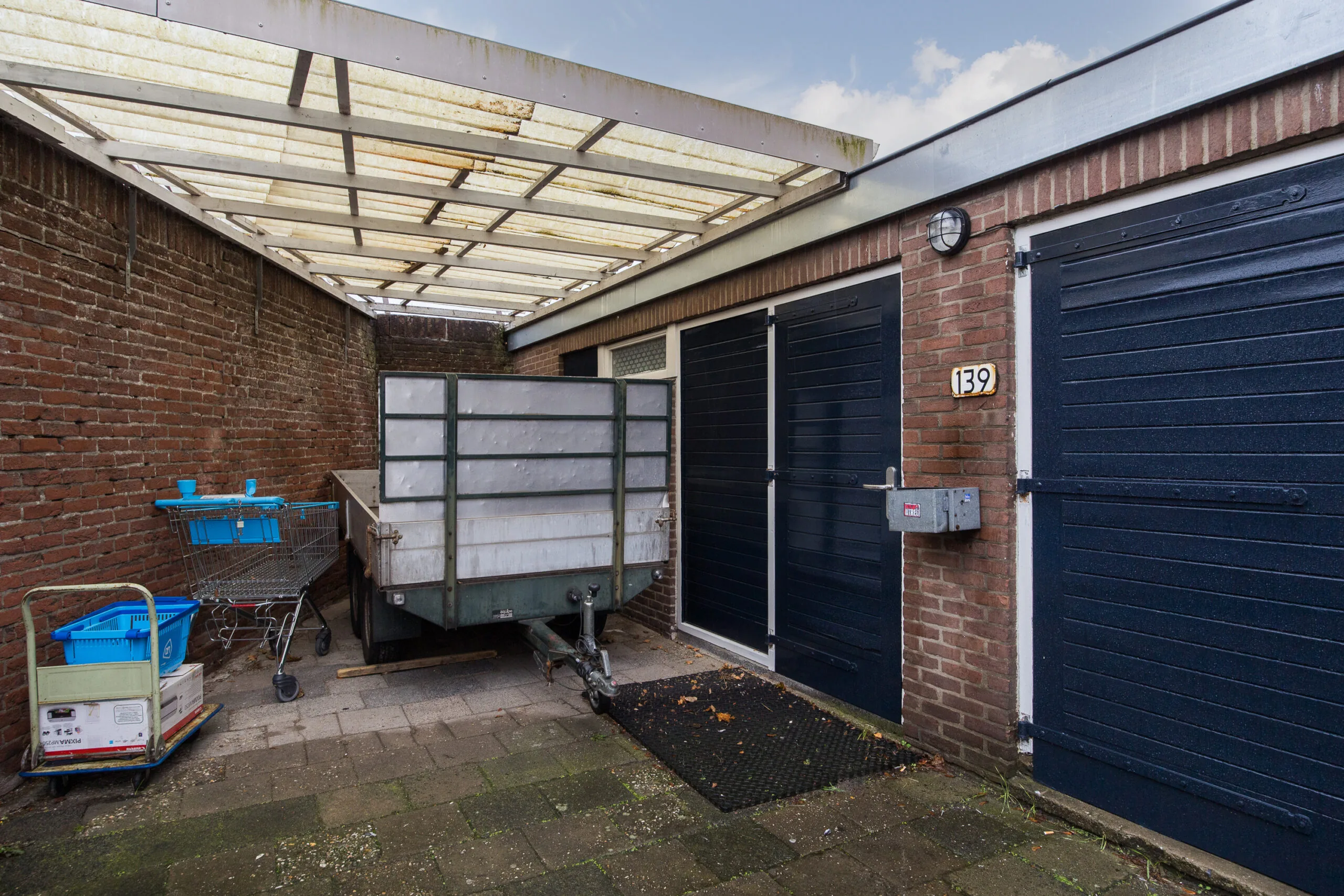 Overdekte steeg met aanhangwagen, winkelwagentje en deur naar huisnummer 139 in de Balderikstraat.