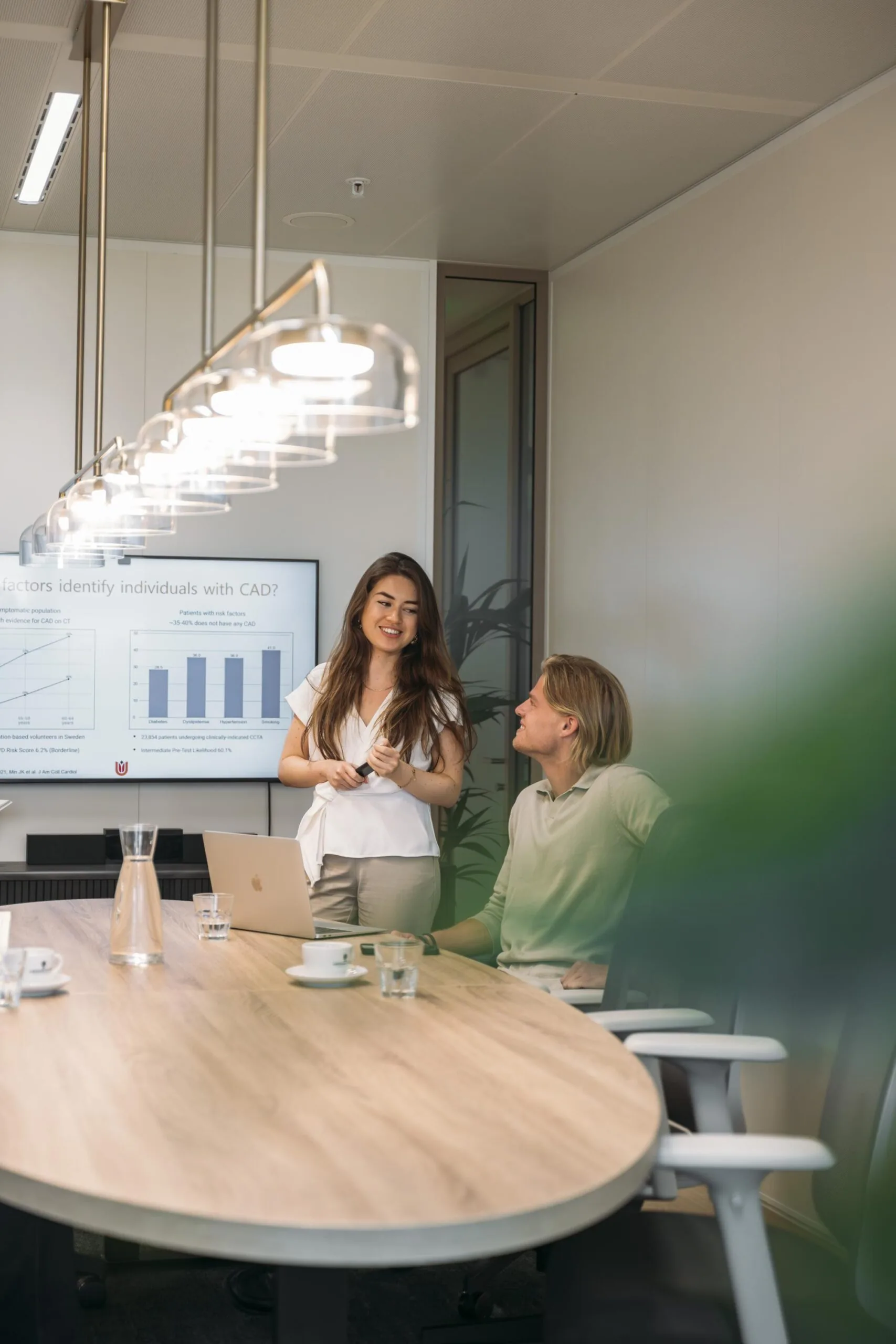 Twee collega's overleggen in een moderne vergaderruimte met presentatie op de achtergrond.