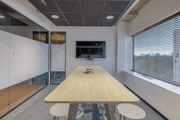 Vergaderruimte met een lange houten tafel, stoelen, een scherm aan de muur en een uitzicht door het raam.