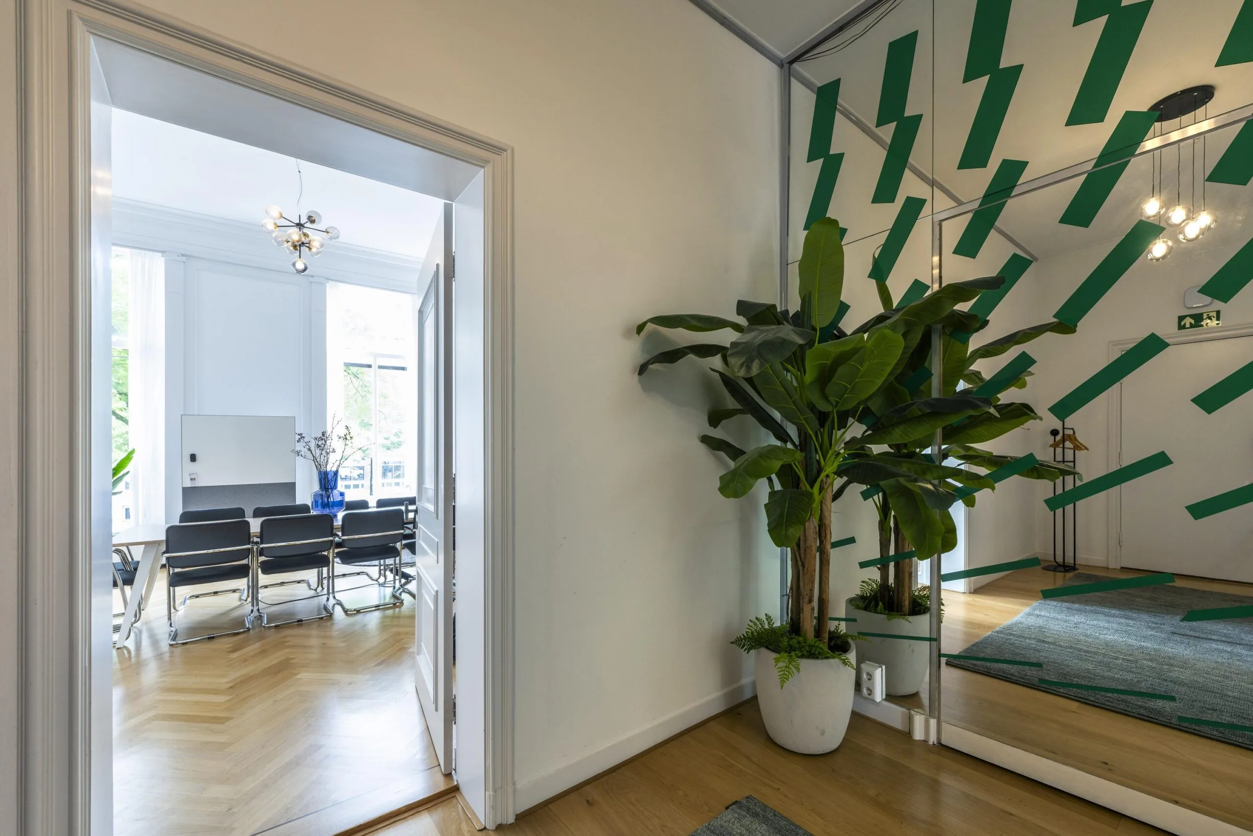 In een modern kantoor aan de Herengracht staan een grote kamerplant, spiegelwand met groene accenten en een vergaderruimte met stoelen en een blauwe vaas op tafel.