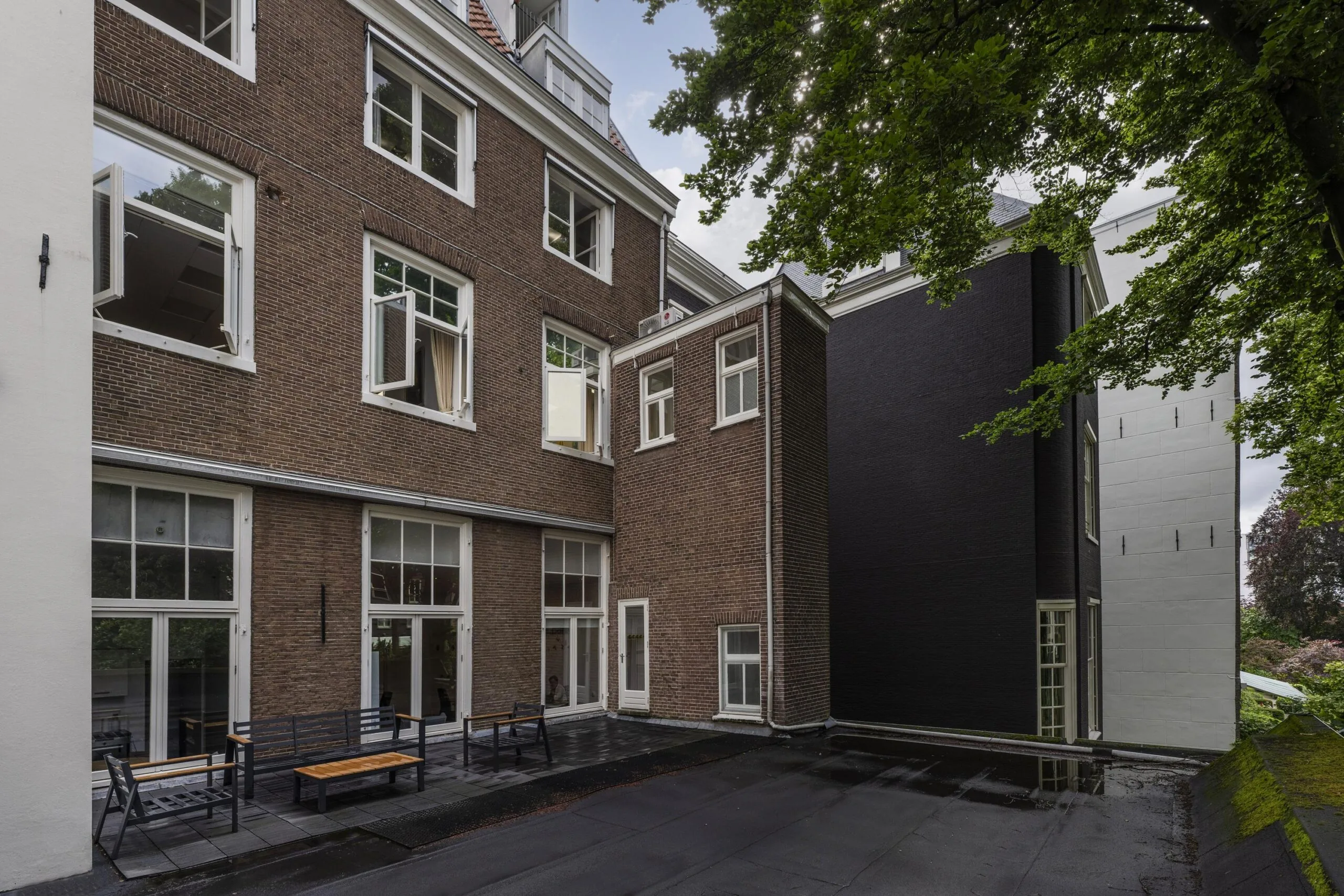 Binnenplaats met zitbanken bij een bakstenen pand aan de Herengracht, omgeven door bomen.