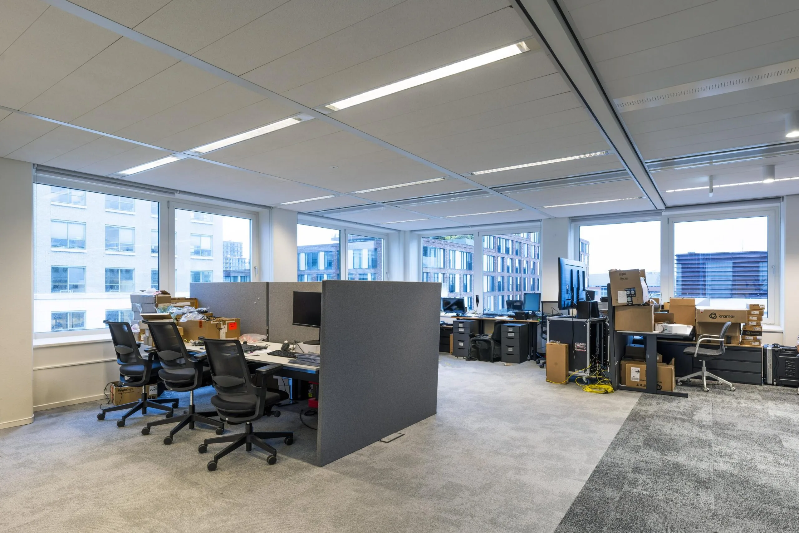 Open kantoorruimte met bureaus, stoelen, computers en stapels dozen in een modern bedrijfsgebouw.