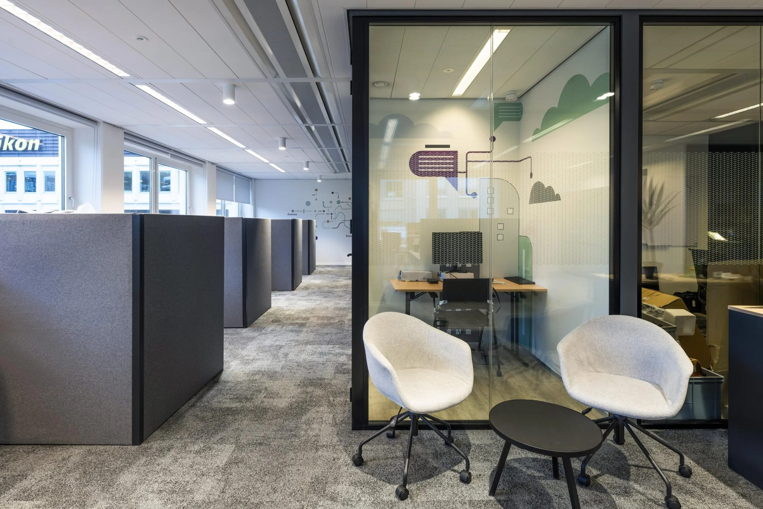 Modern ingericht kantoor met werkplekken, glazen vergaderruimte en wachthoek met stoelen en tafel.