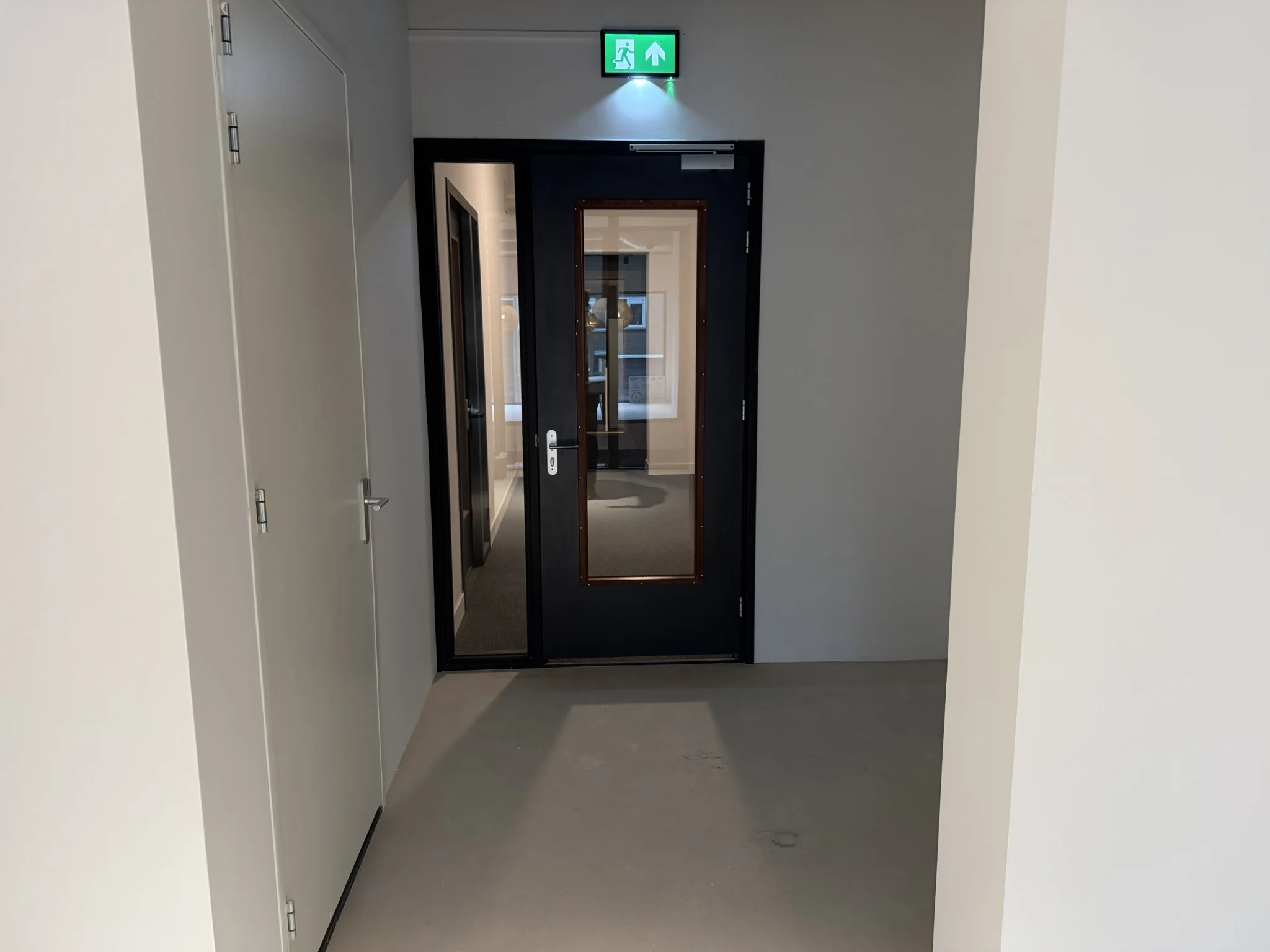 Hal met grijze vloer en witte muren, leidend naar een zwarte deur met glazen paneel en groen nooduitgangsbord erboven.