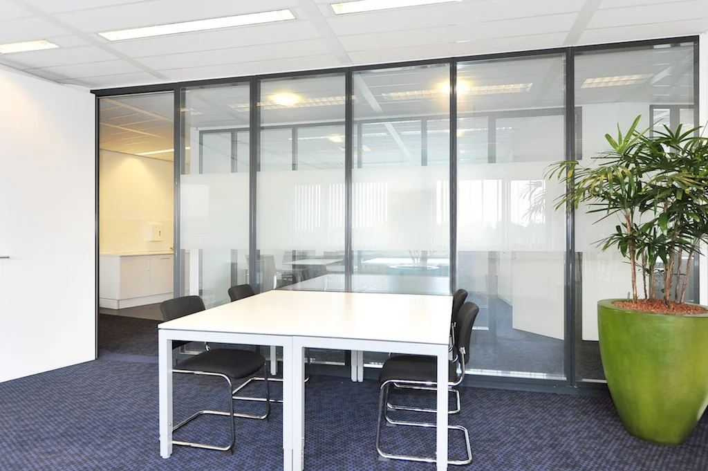Moderne vergaderruimte aan de Laan van ’s-Gravenmade met glazen wanden, witte tafel, zwarte stoelen en een grote kamerplant.