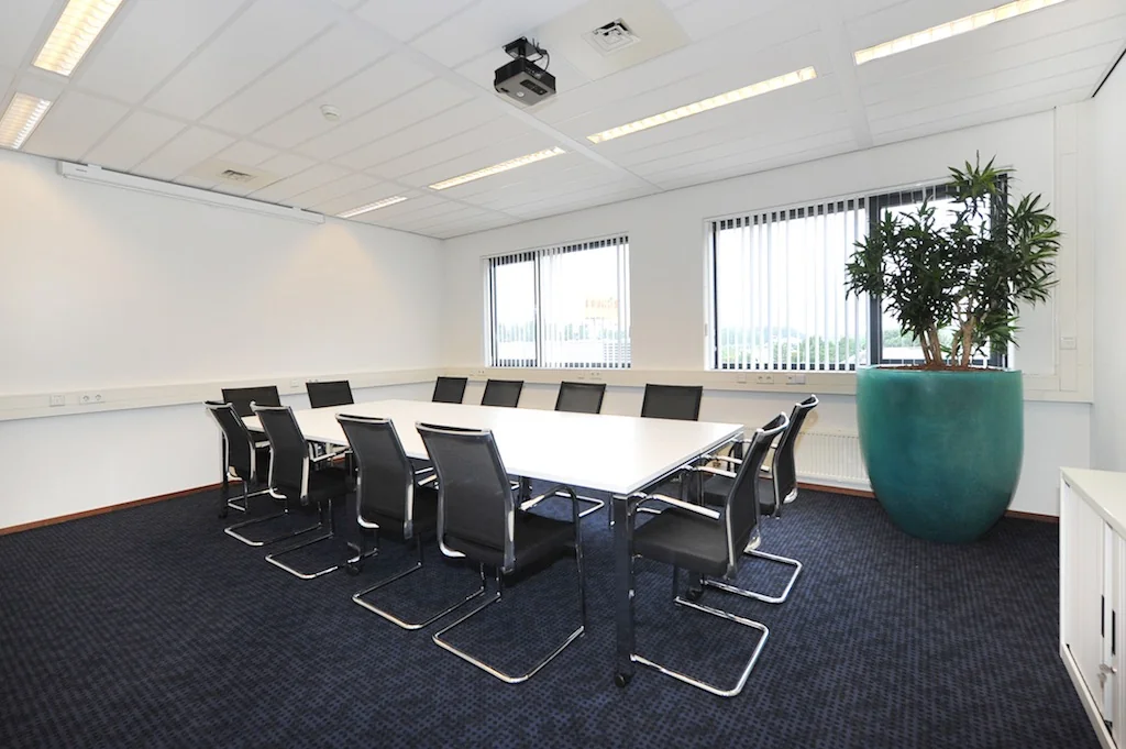 Vergaderruimte aan de Laan van ’s-Gravenmade met een lange tafel, zwarte stoelen, grote plantenbak en een projector aan het plafond.